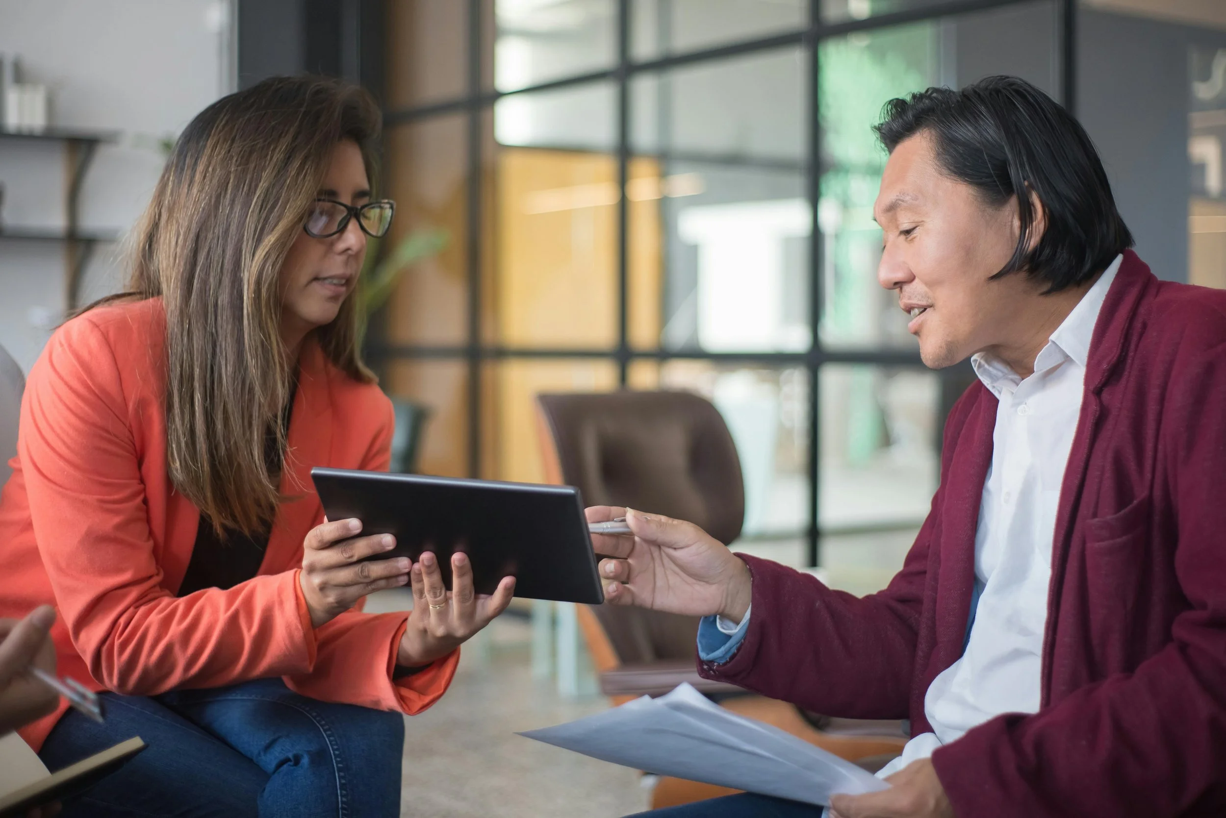 A man in a suit jacket points to something on a digital tablet held by a woman in a suit jacket. They are sitting in an office, coordinating on a project.