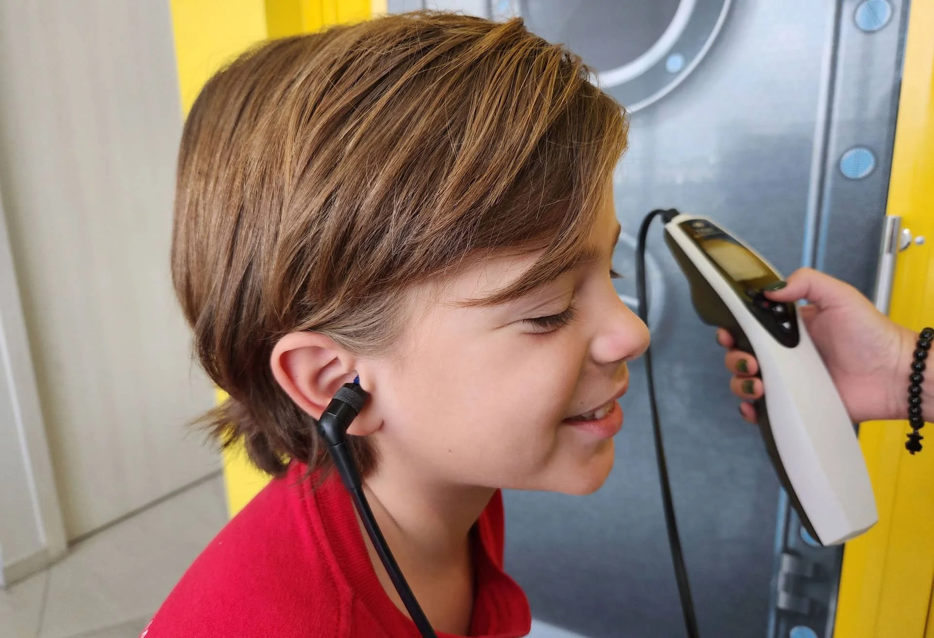Una niña sonriendo, siendo sometida a un examen de temperatura con un termómetro digital por una persona cuyas manos se ven, en un entorno que parece ser una escuela o clínica.