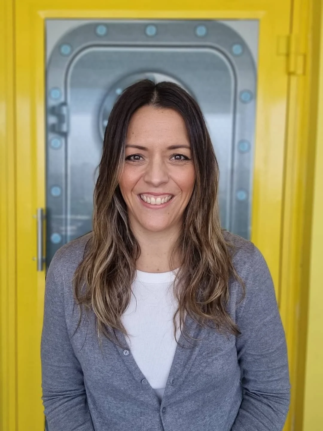 Retrato de una mujer con cabello largo y ondulado, sonriendo, con fondo de una puerta metálica de color gris y marco amarillo.