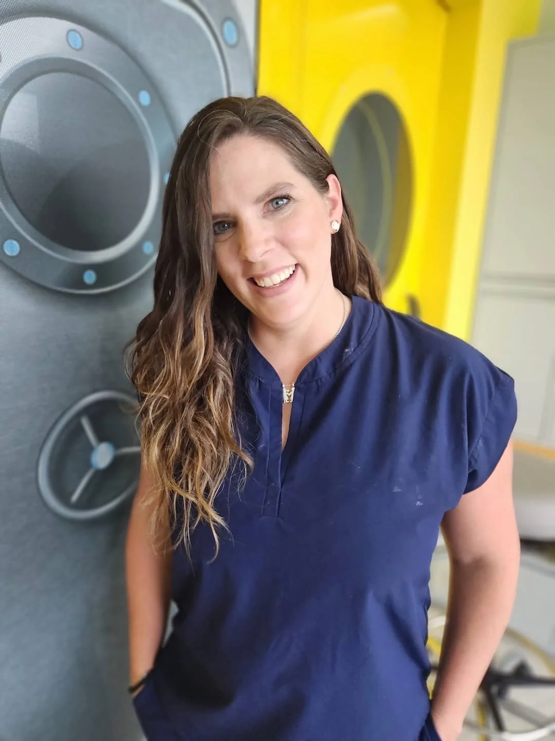 Mujer sonriendo con cabello rizado, usando uniforme azul, en lavandería con lavadoras amarillas y gris oscuro.