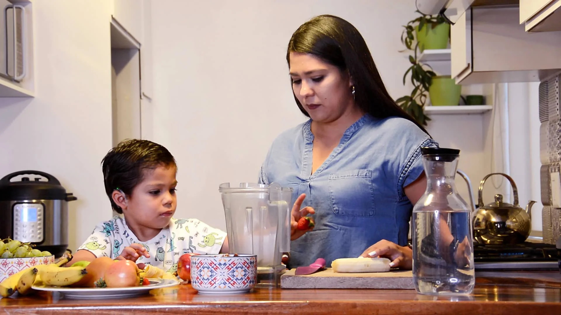 Una mujer y un niño en la cocina preparando jugo con frutas variadas, como plátanos, uvas, manzanas y fresa.
