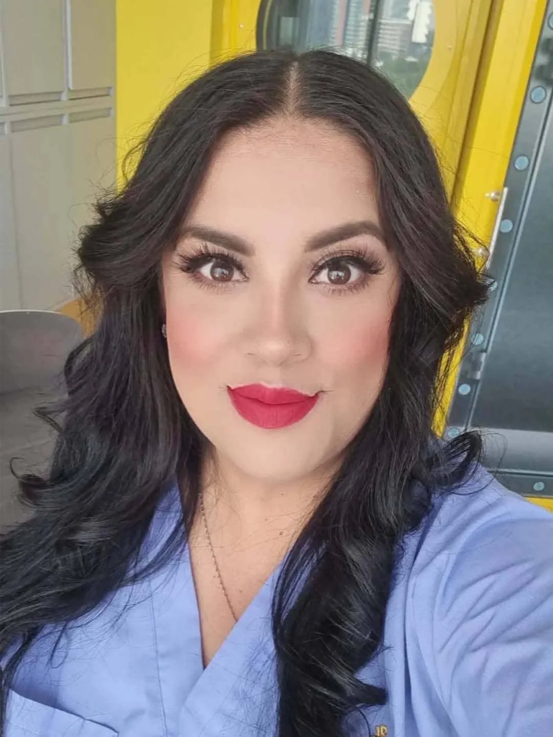 Mujer con cabello rizado oscuro, maquillaje detallado y ropa azul, sonriendo en un entorno industrial.