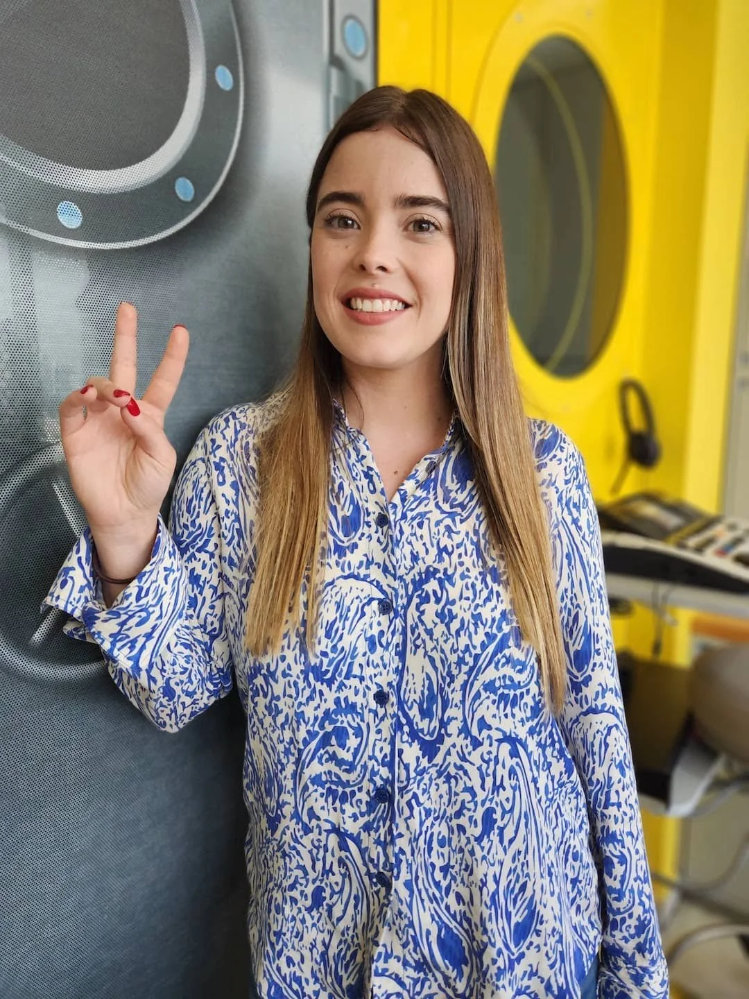 Joven mujer sonriendo y haciendo señal de paz con el dedo índice y medio en una lavandería con lavadoras amarillas.