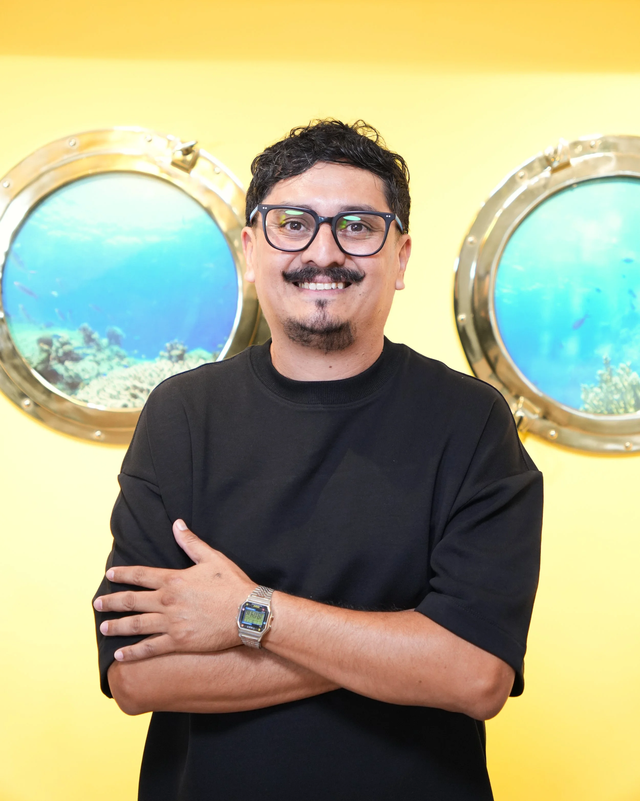 Hombre con bigote y camiseta negra con dibujo de personajes de caricatura, sonriendo y haciendo pulgar arriba, en una cabina de grabación o estudio de música.