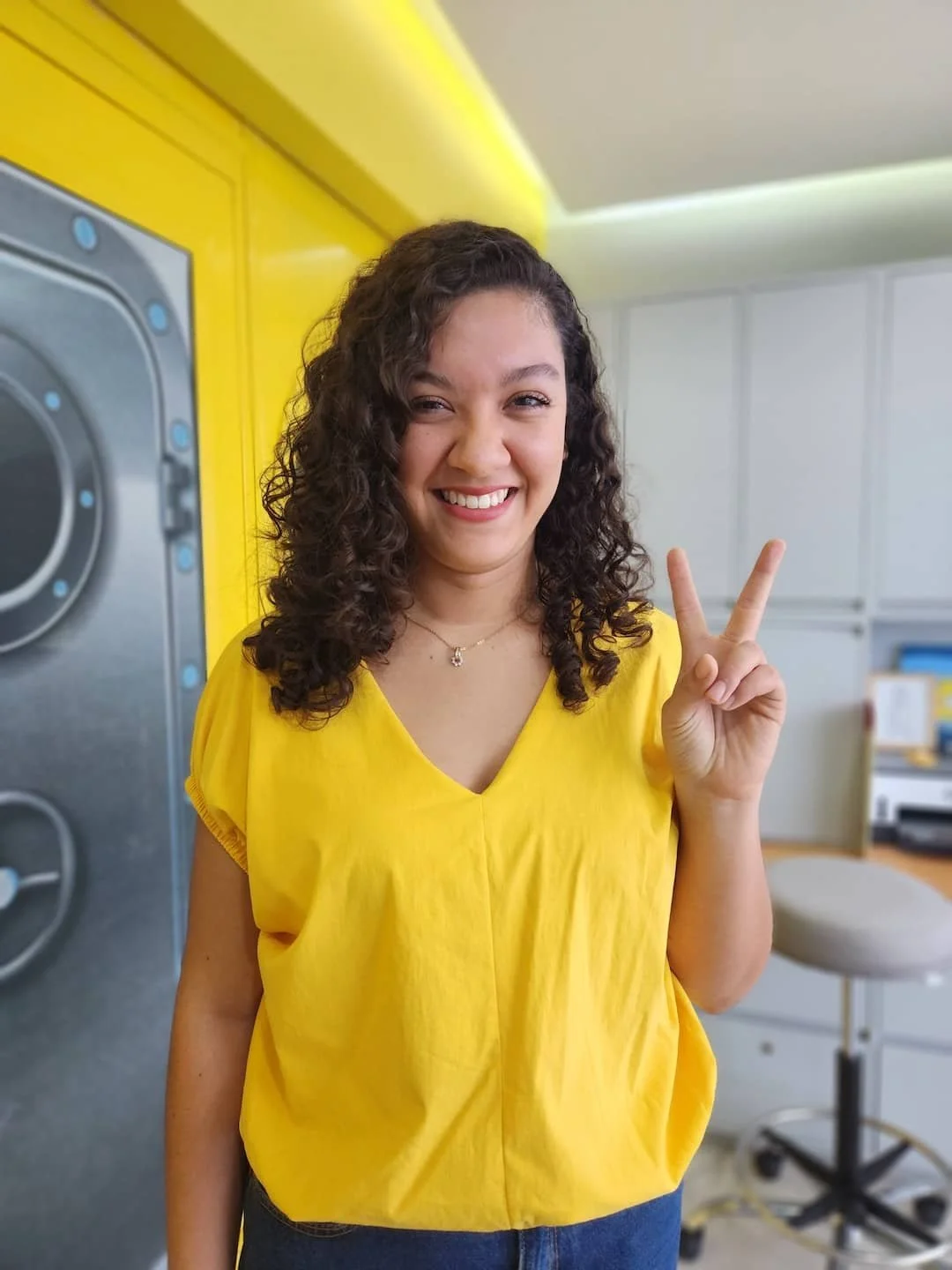 Una joven sonriente con cabello rizado, vistiendo una blusa amarilla y haciendo una señal de paz con la mano, en un entorno de lavandería.