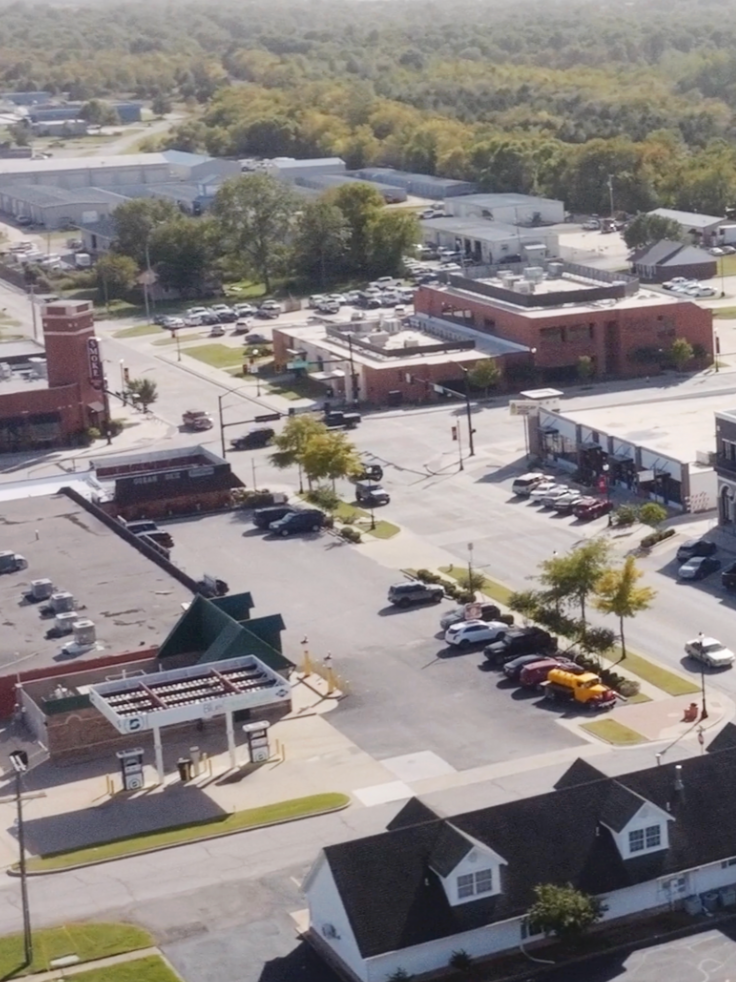 Aerial view of a small town with a parking lot, streets, retail stores, and surrounding greenery.