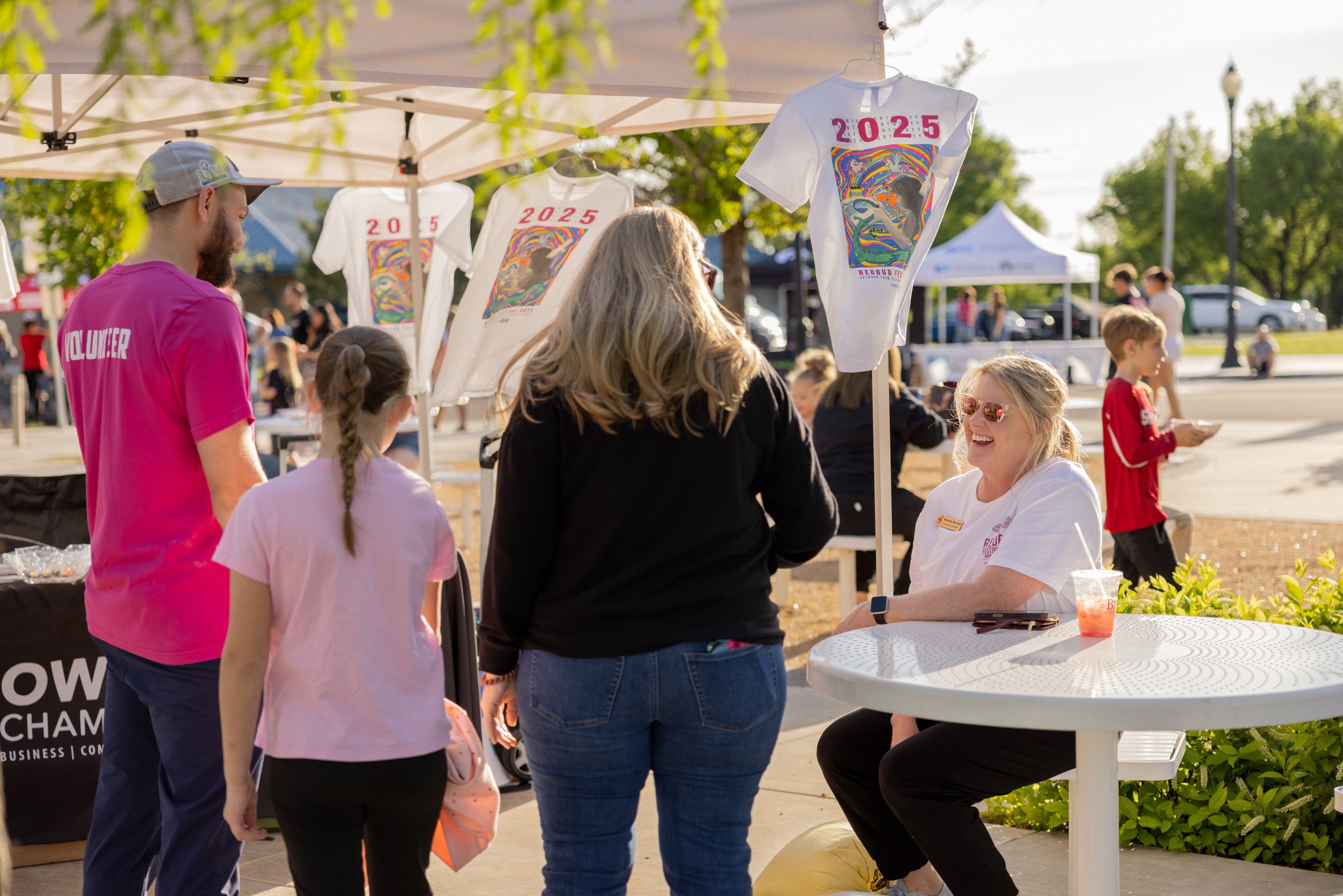 A woman with blonde hair and sunglasses sitting at a white round table, smiling and talking to a woman with gray hair in front of her. Two children with braided hair are also facing the woman with gray hair. Behind them, a man in a pink shirt with volunteer printed on the back, and T-shirts with the year 2025 are displayed at a booth outdoor event on a sunny day.