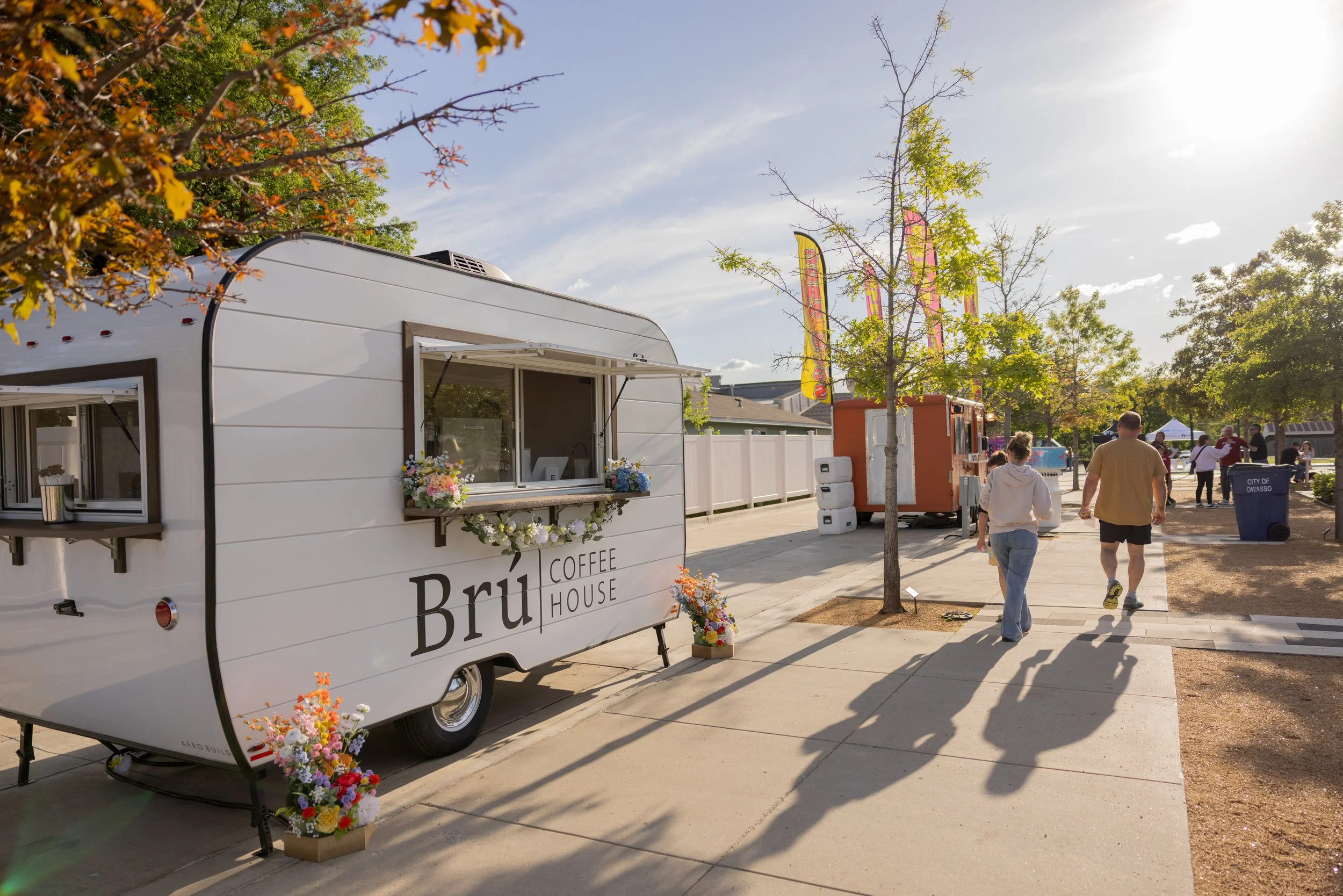 A white coffee truck with floral decorations parked on a sidewalk at a fair or outdoor event. People are walking along the sidewalk, and there are trees, flags, and event tents in the background.