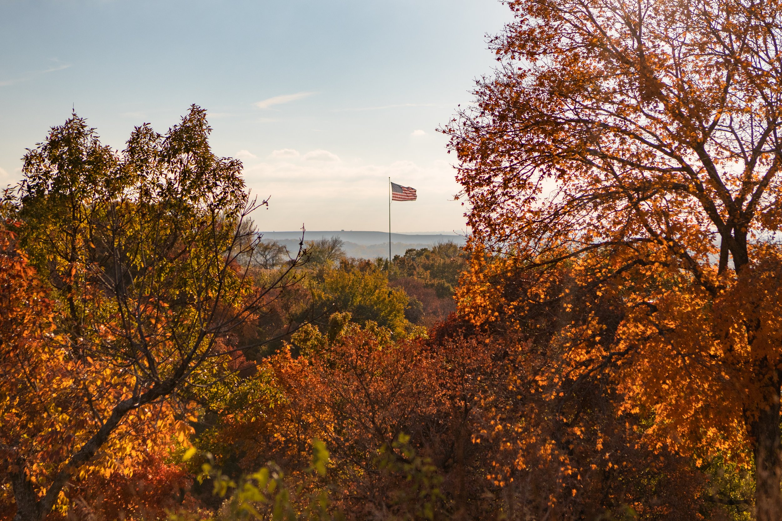 Autumn trees with colorful fall foliage and an American flag flying in the distance under a partly cloudy sky.