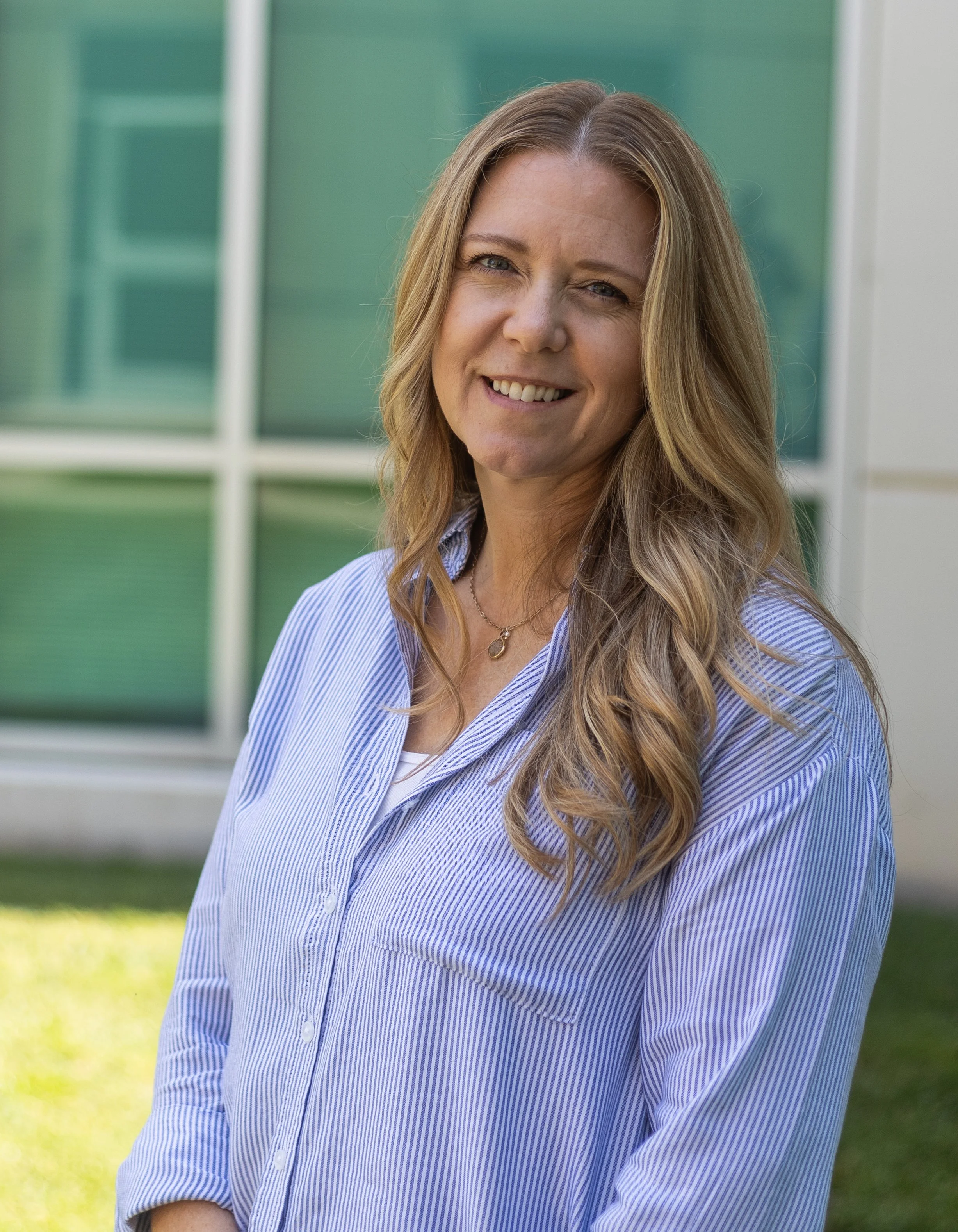 A woman with blonde, wavy hair smiling outdoors in front of a modern glass building, wearing a light blue and white striped shirt and a necklace.