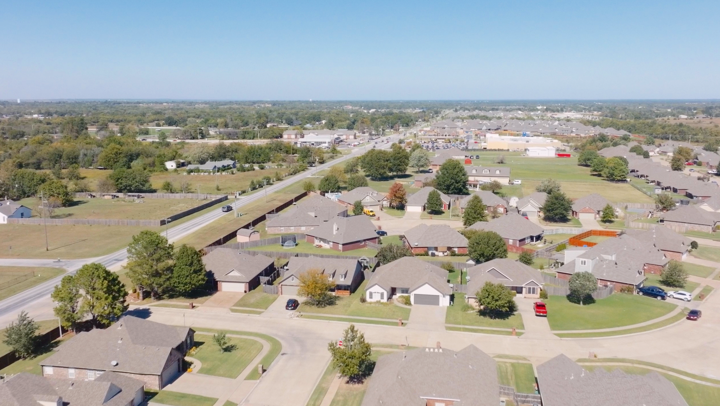 Aerial view of a suburban neighborhood with houses, trees, roads, and a commercial shopping area in the background on a clear day.