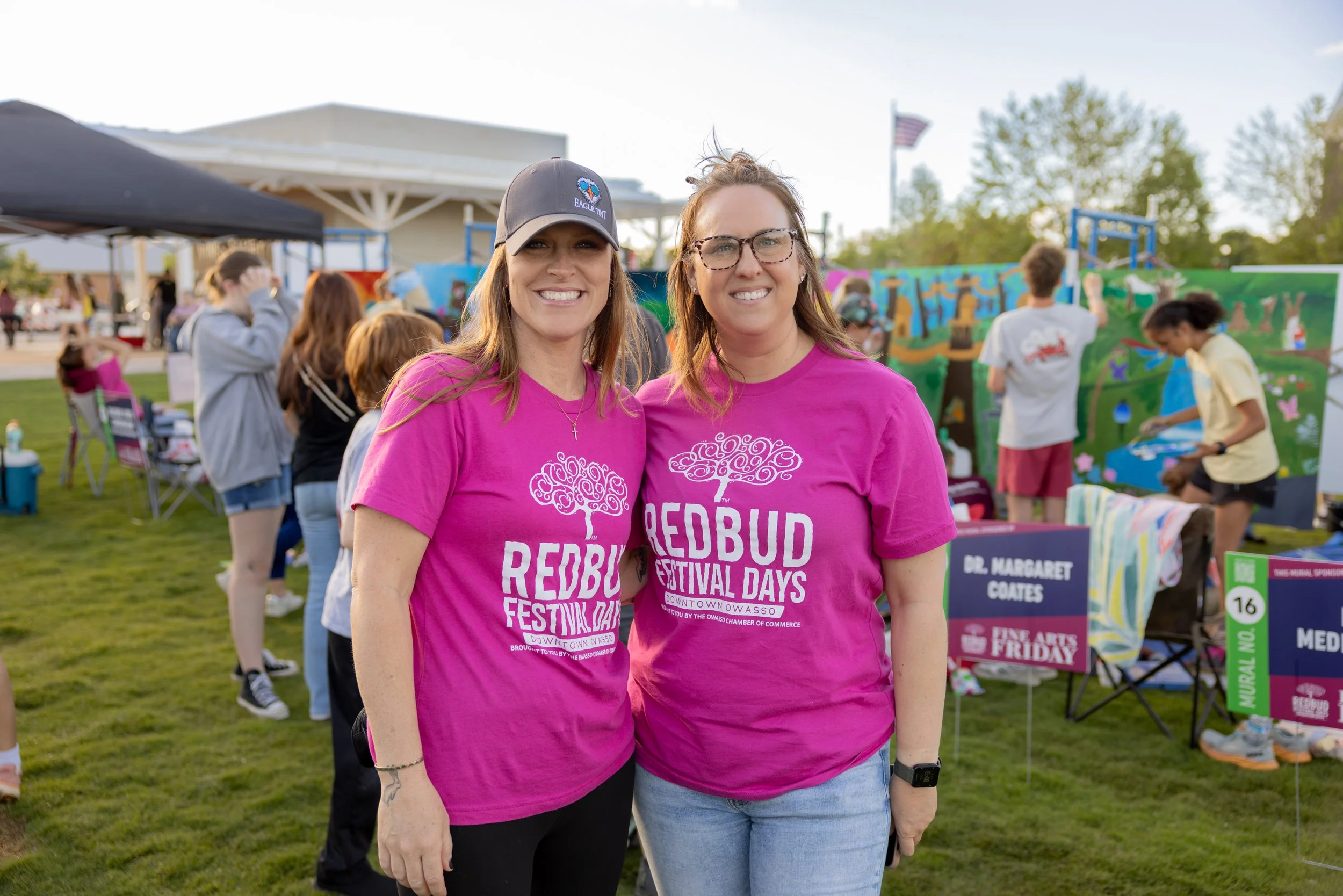Two women smiling at the camera during an outdoor festival, wearing matching bright pink T-shirts with the text "Redbud Festival Days" and a tree logo, with people and art displays in the background.