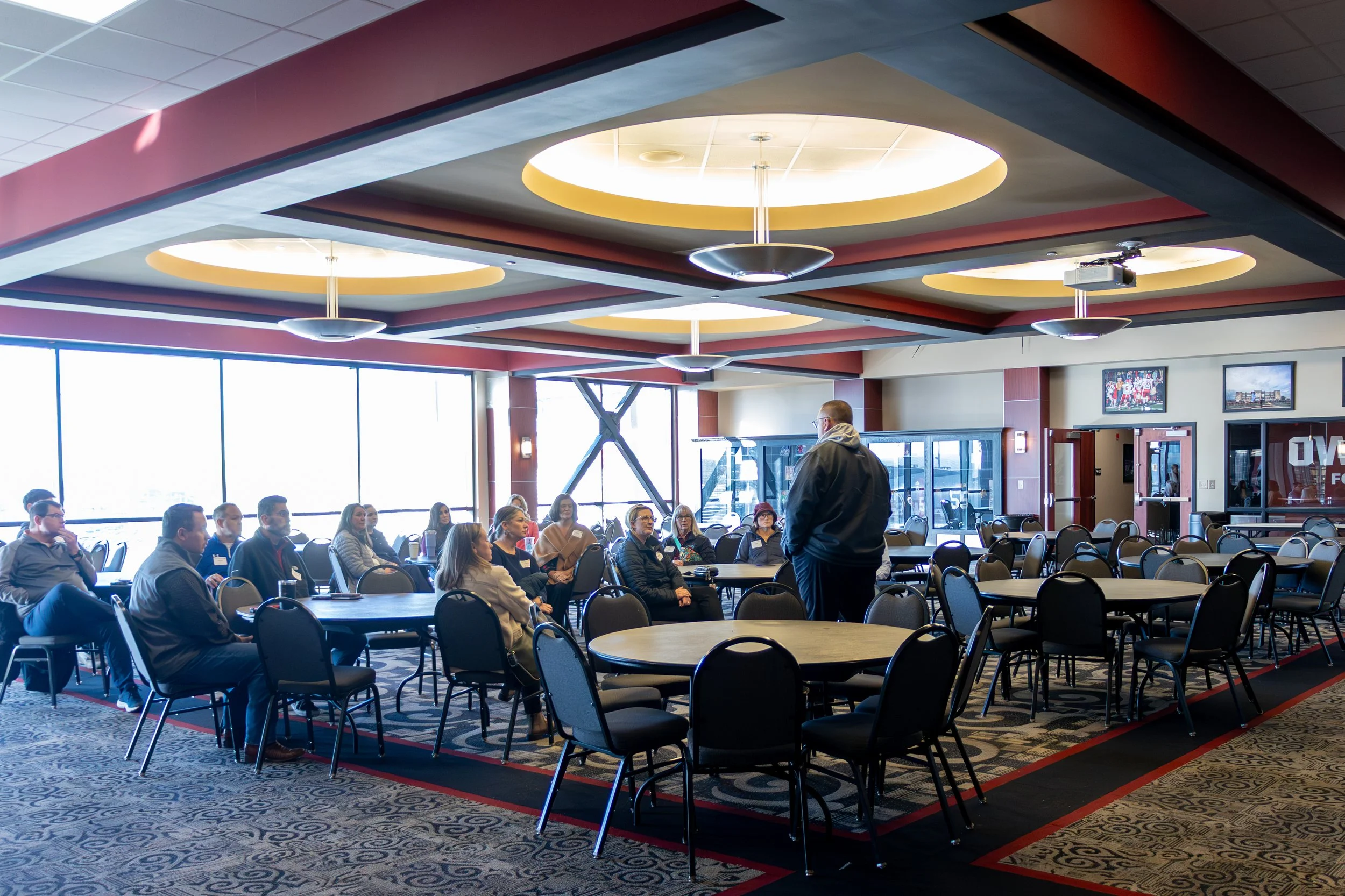 A man standing and speaking to a group of people seated at round tables in a conference room.