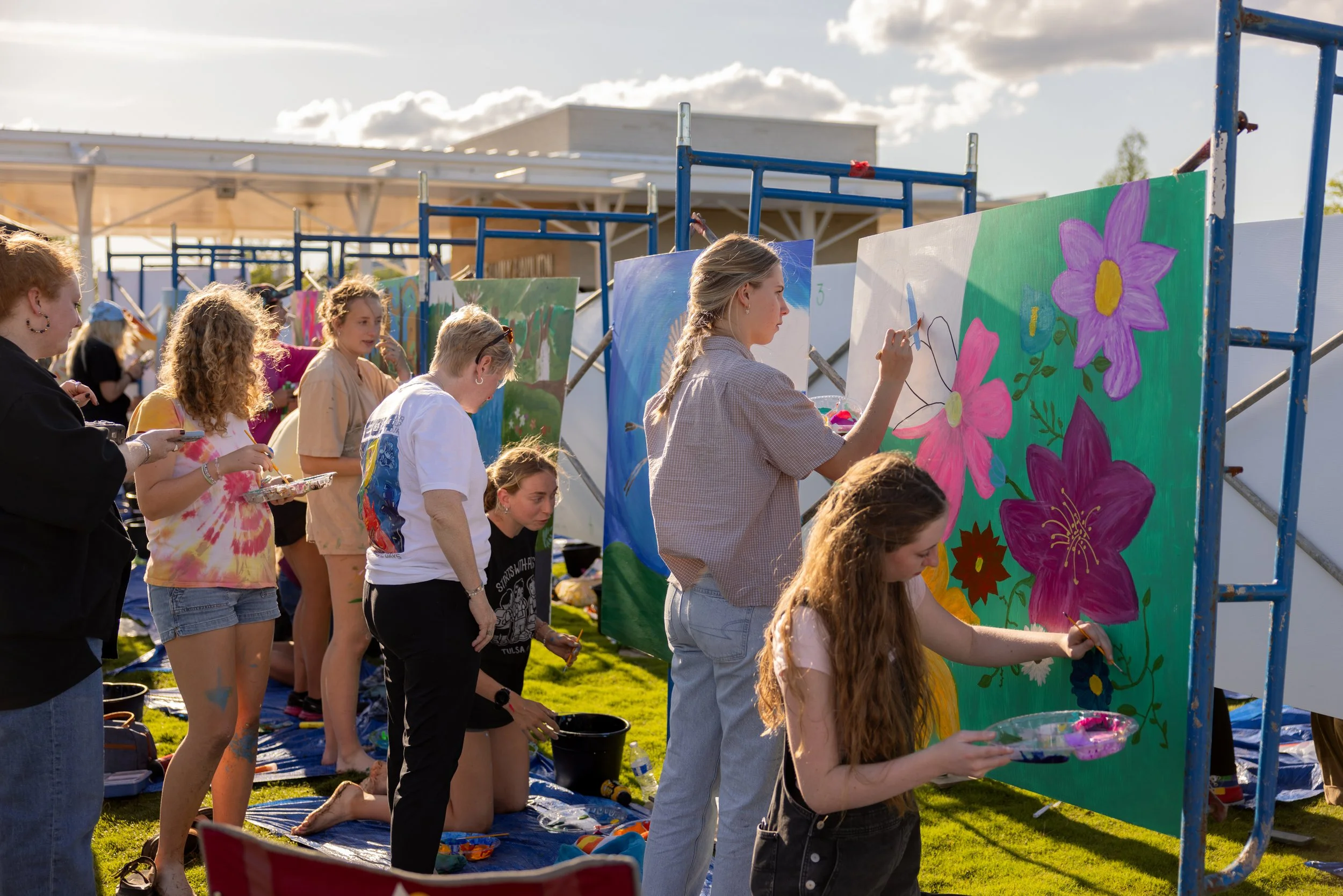 People painting large floral murals on outdoor panels at an art event on a grassy area.