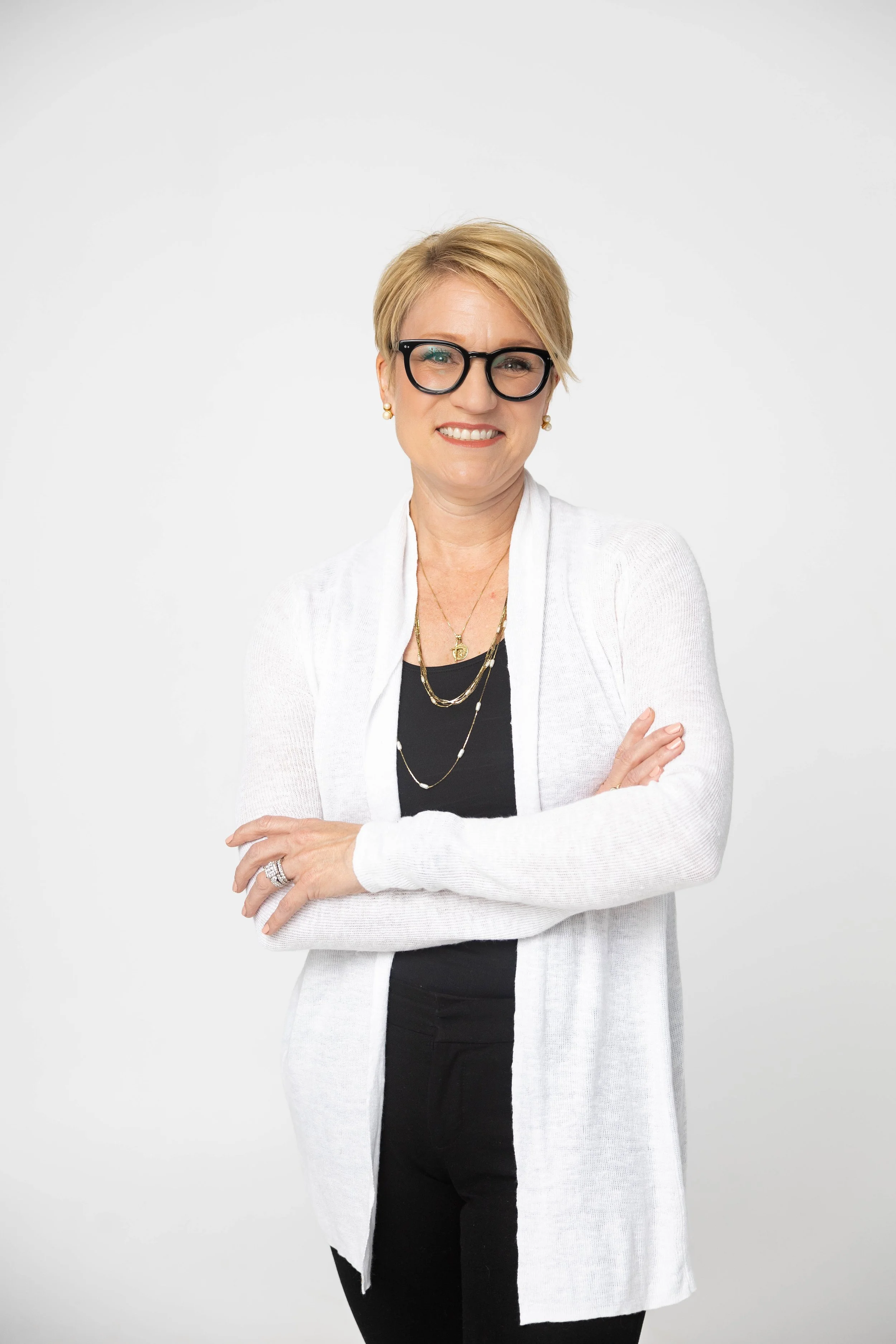 A middle-aged woman with short blonde hair, wearing glasses, a white cardigan, black top, and black pants, standing with arms crossed and smiling against a plain white background.