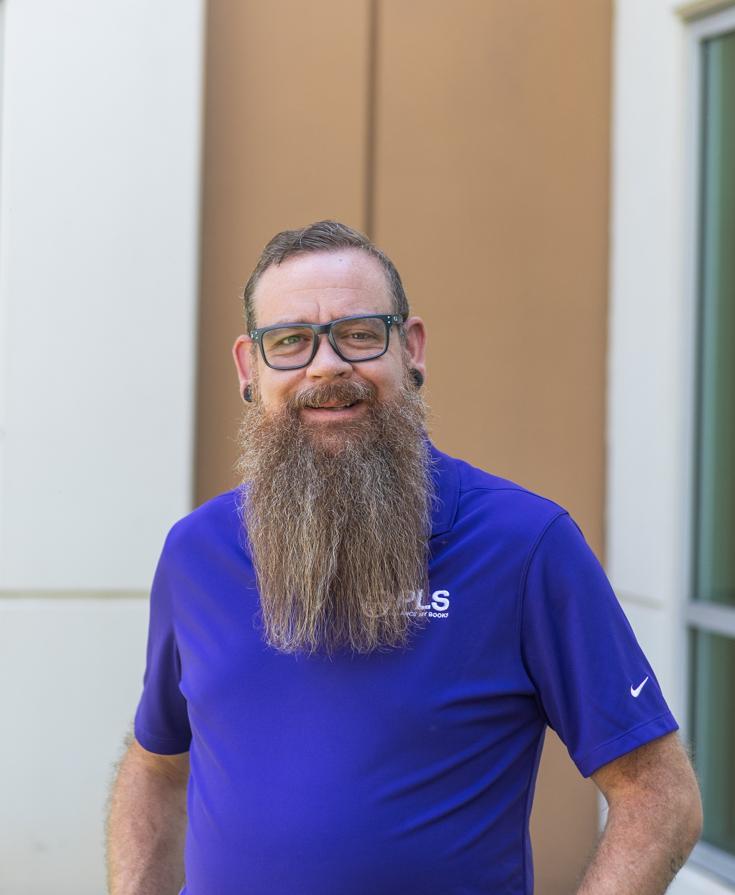A man with glasses and a long beard wearing a blue Nike sports shirt, standing outside near a building.