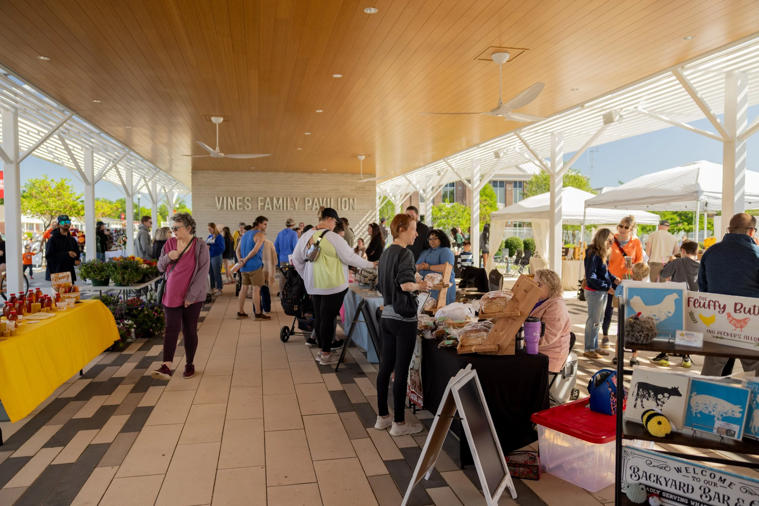 People shopping at outdoor vendor booths under a covered pavilion labeled 'Vines Family Pavilion' with white tent canopies and a wooden ceiling, during a market event.