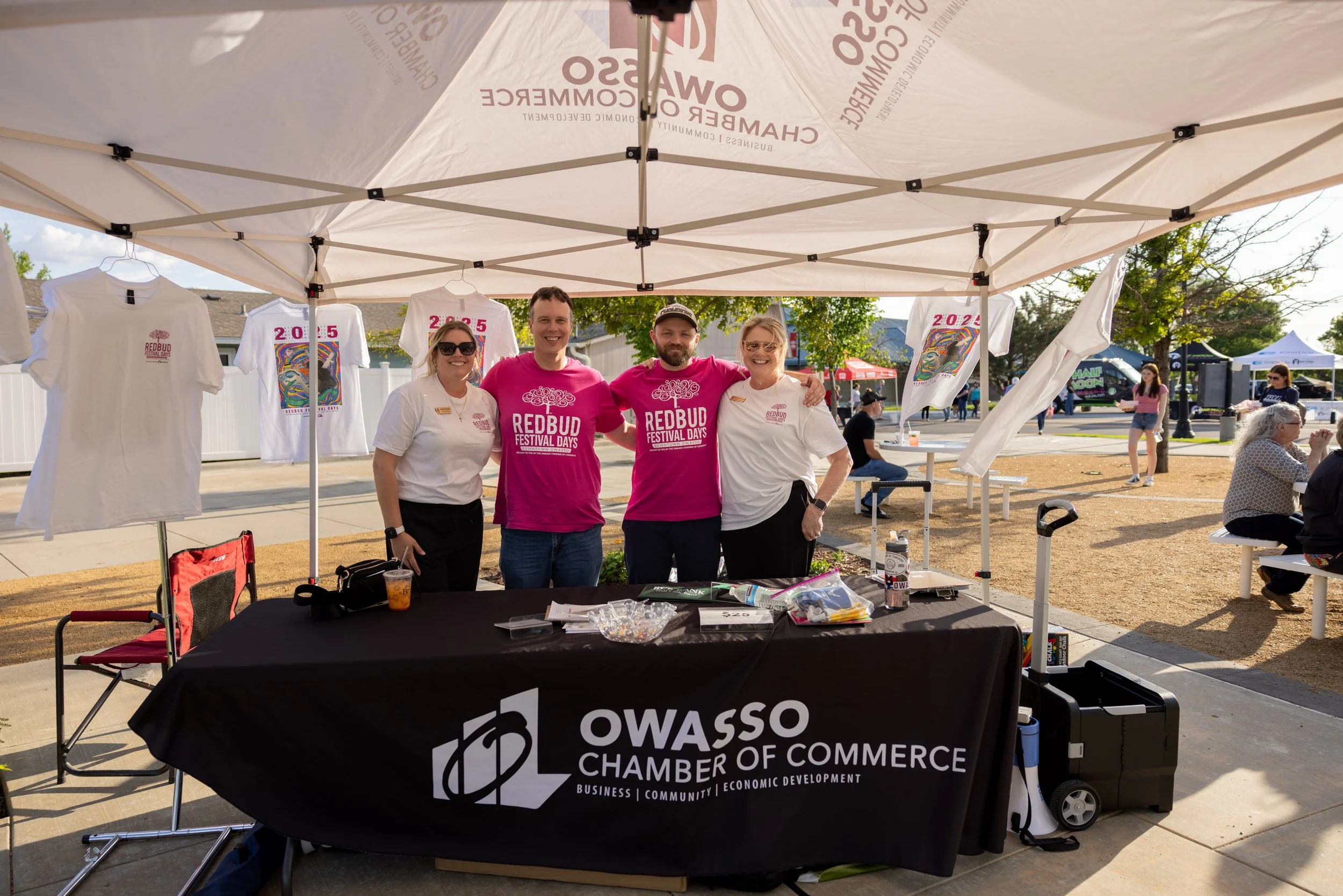 Four people standing behind a table at an outdoor event under a white canopy tent. The table has a black cloth with the Owasso Chamber of Commerce logo. Two women and two men are smiling and posing for the camera. The women are wearing white shirts and the men are wearing bright pink shirts with 'Redbud Festival Days' printed on them. T-shirts with event graphics are hanging on the tent. There are other people and additional festival tents in the background.