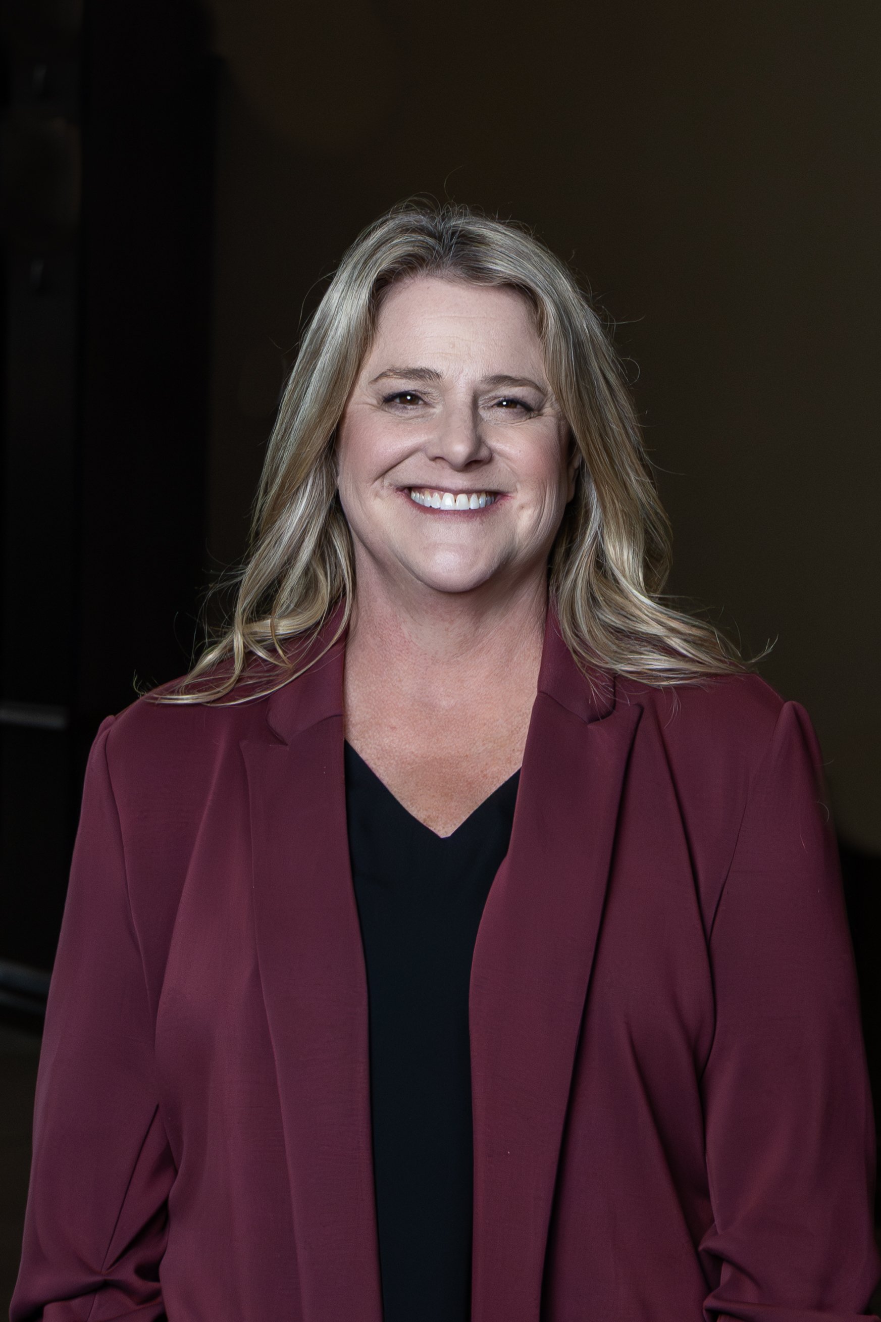A smiling woman with long blonde hair wearing a maroon blazer and black top against a dark background.