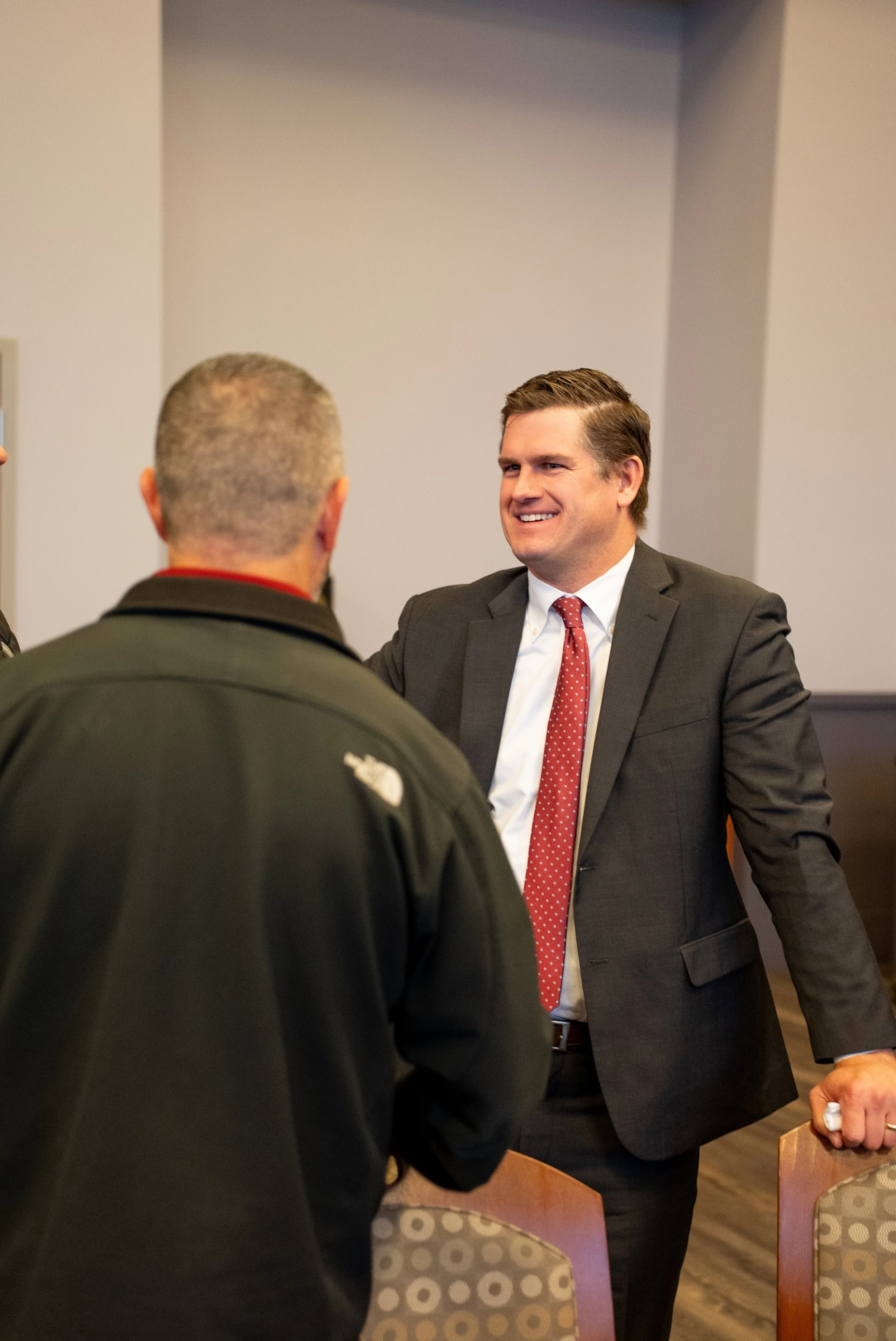 Three men engaged in conversation, with one smiling and dressed in a black suit with a red tie, while the other two are in dark jackets, all standing in a room with wooden chairs and a plain wall.