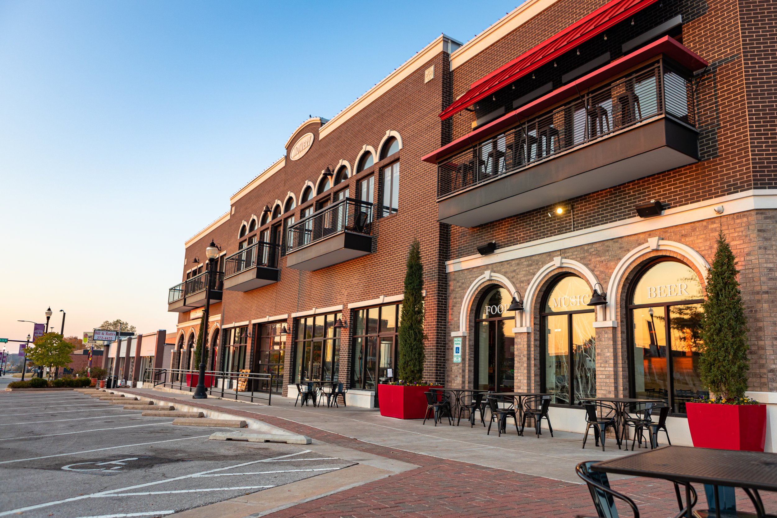 Exterior view of a brick commercial building with large windows and outdoor seating, under a clear sky, with parking spaces in front.