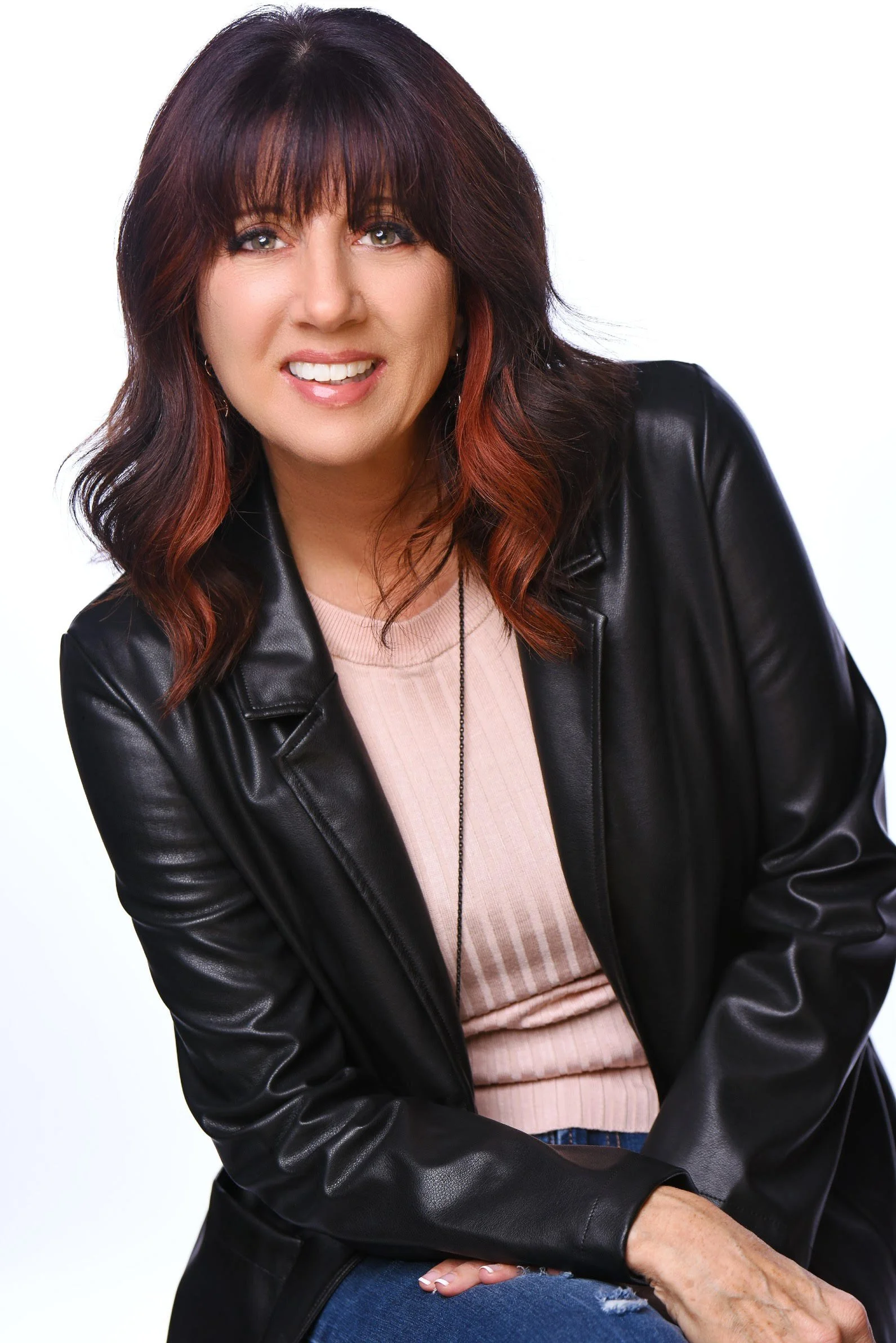 A woman with shoulder-length, dark brown hair with red highlights, wearing a black leather jacket over a beige top, smiling at the camera against a plain white background.