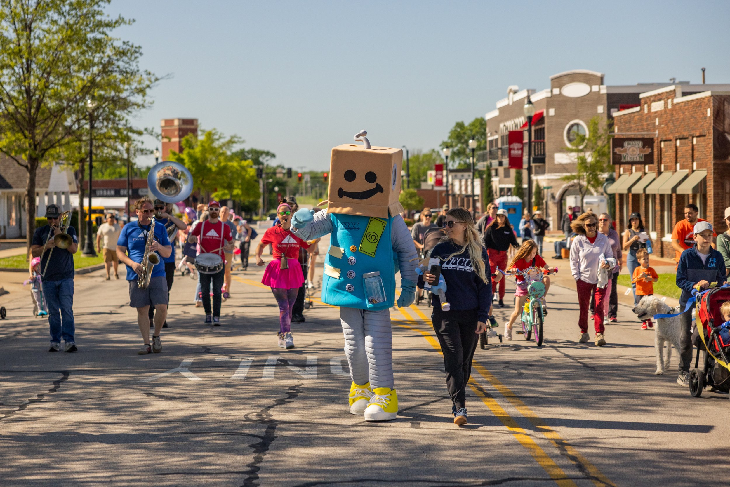 A person dressed in a robot costume walking with a young girl on a street during a parade or event, surrounded by people playing musical instruments, children, and spectators. The street has painted parking lines and is lined with trees and buildings.