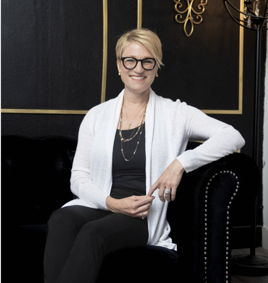 A woman with short blonde hair, wearing glasses, a white cardigan, and a black outfit, sitting on a black velvet couch with decorative gold accents in the background.