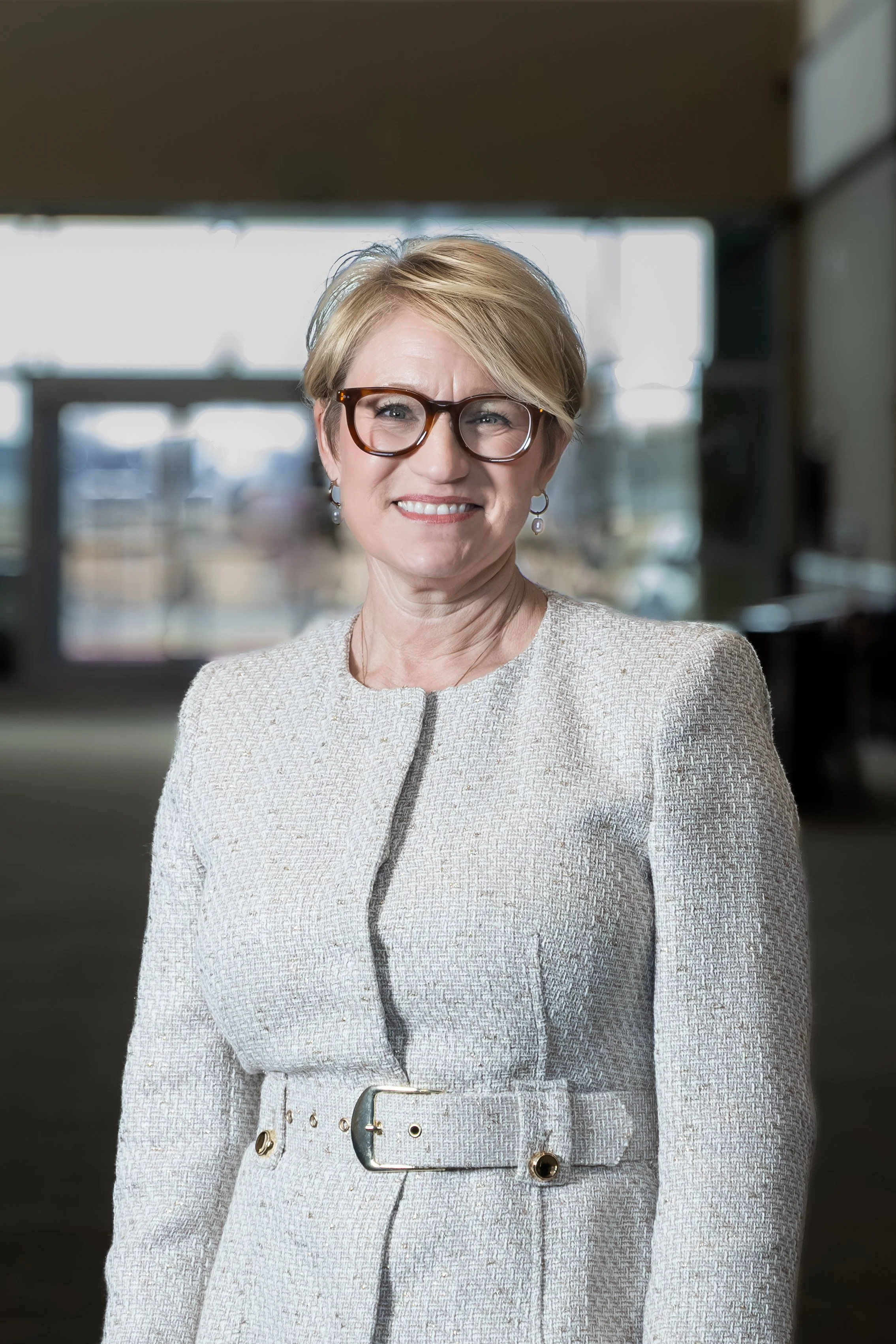 A smiling middle-aged woman with short blonde hair and glasses, wearing a light-colored textured dress with a belt, standing indoors in front of windows and a blurry background.