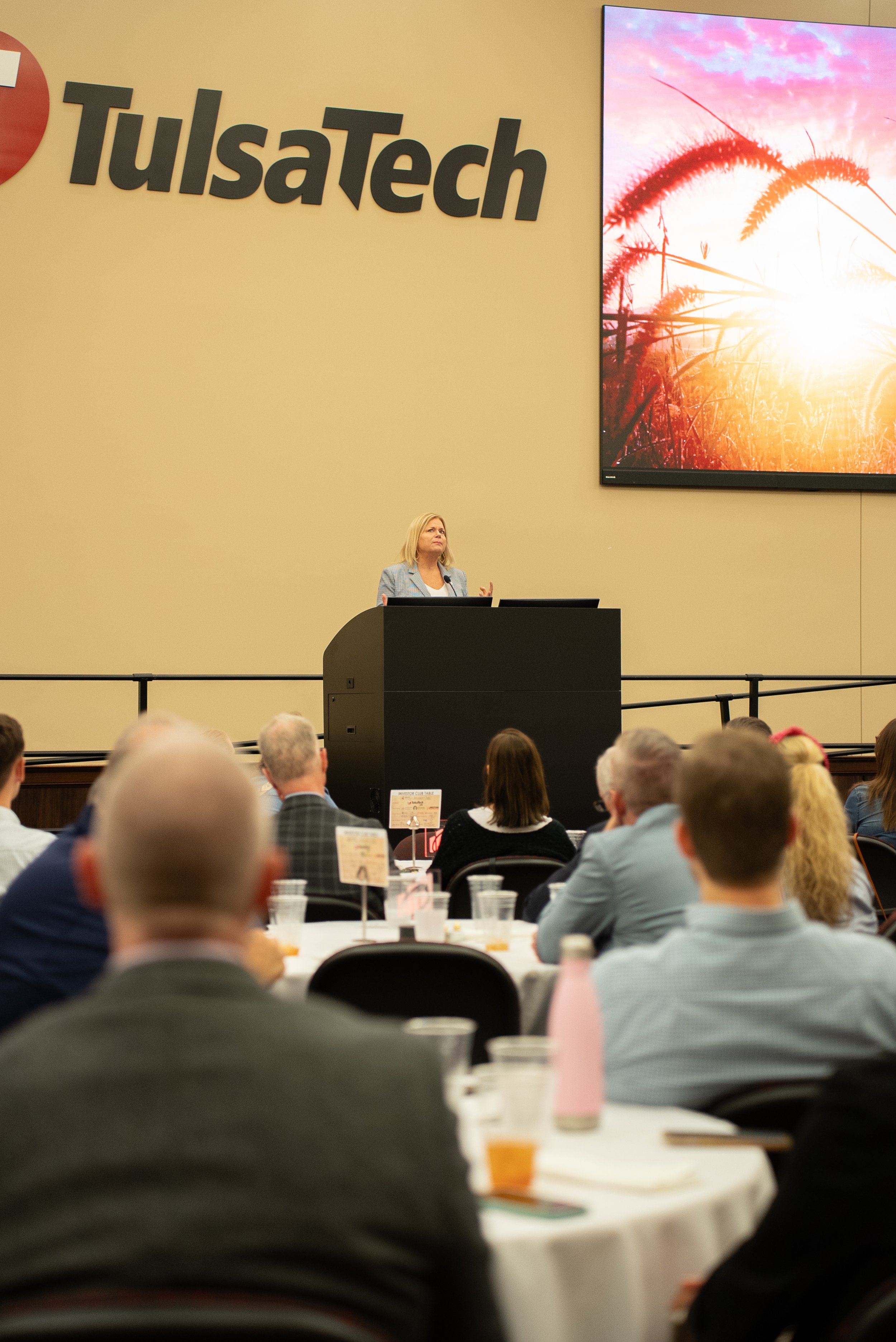 A woman giving a presentation at Tulsatech conference, with a large screen displaying an image of a sunrise over grassy fields and a crowded audience seated at round tables.