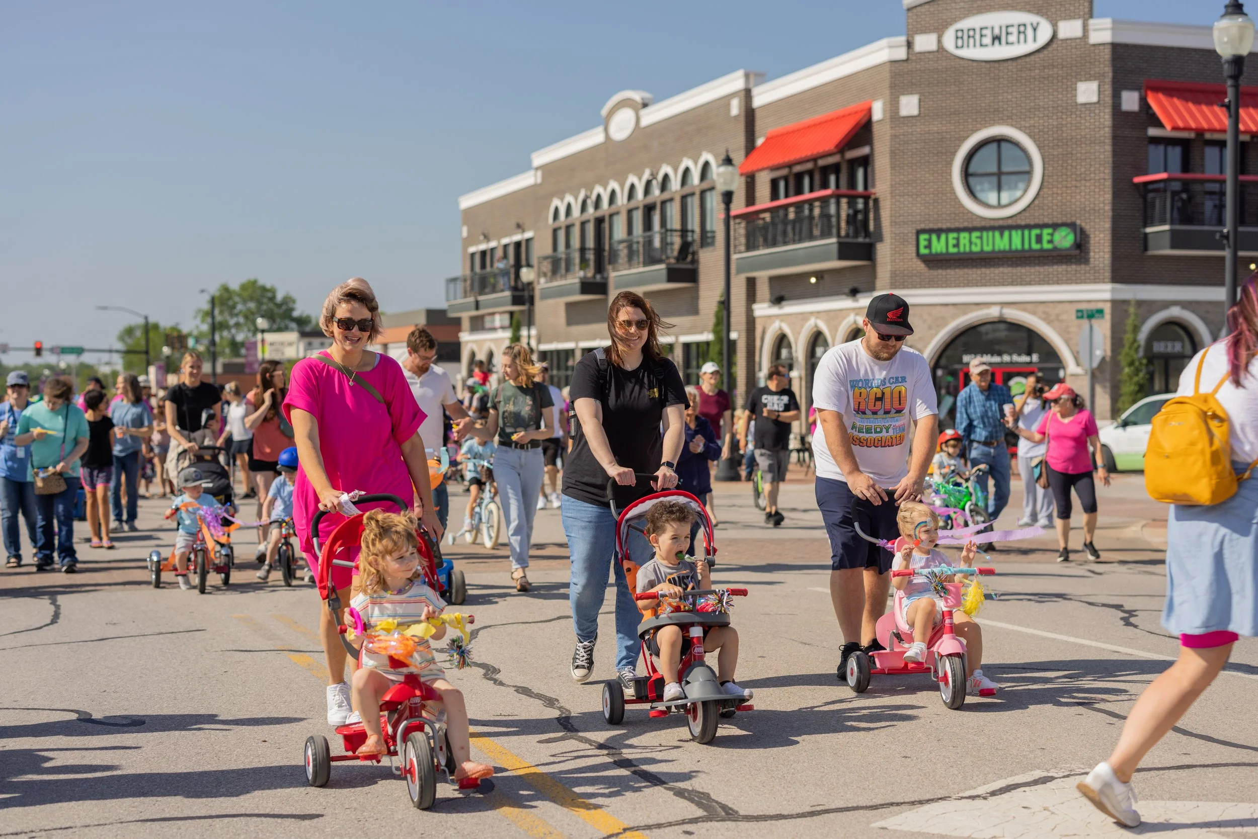People participating in a community bike and stroller parade on a city street, with children riding small bikes and tricycles, and adults walking alongside. The background shows a shopping area with a brewery and a shop named EmerSumIce.
