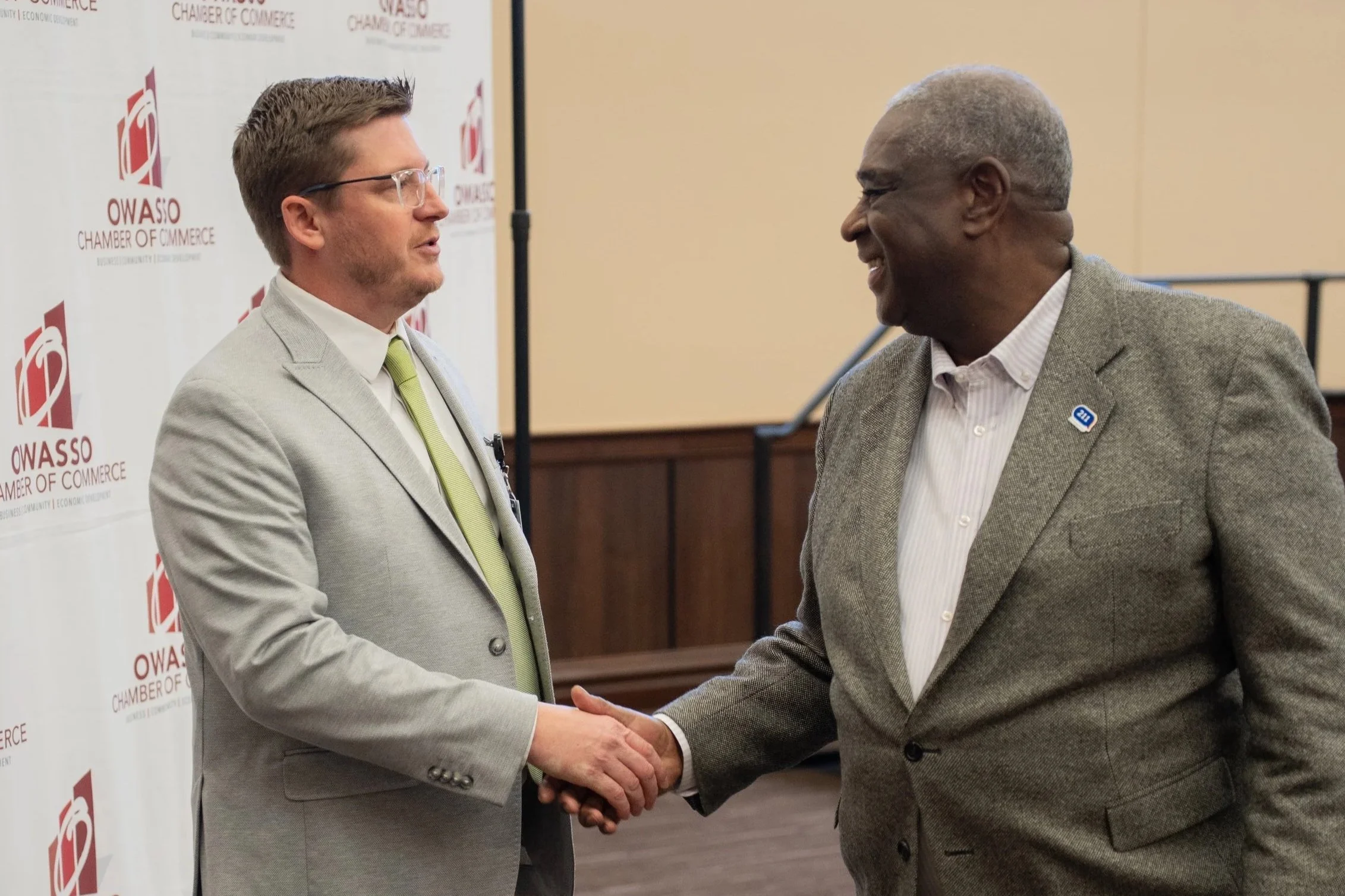Two men shaking hands at a formal event, standing in front of a backdrop with OWASSO Chamber of Commerce logo.