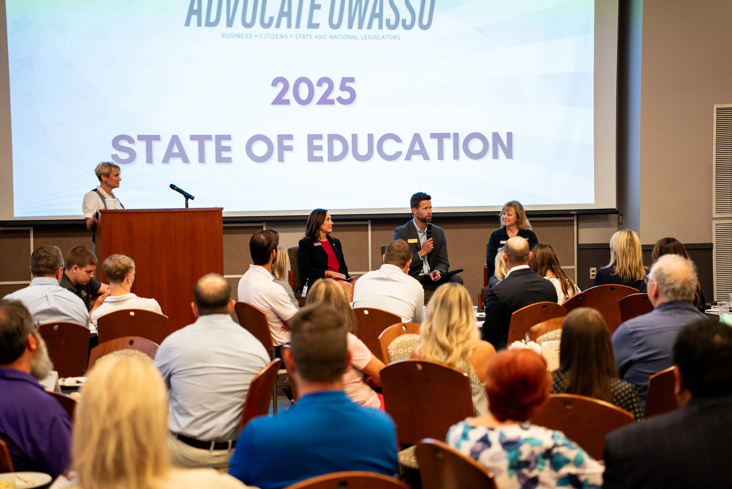 A woman standing at a podium speaking to an audience at the 2025 State of Education event, with three panelists seated on stage in front of a large screen that reads 'Advocate Owasso 2025 State of Education' and 'Business + Citizens + State and National Legislators.'