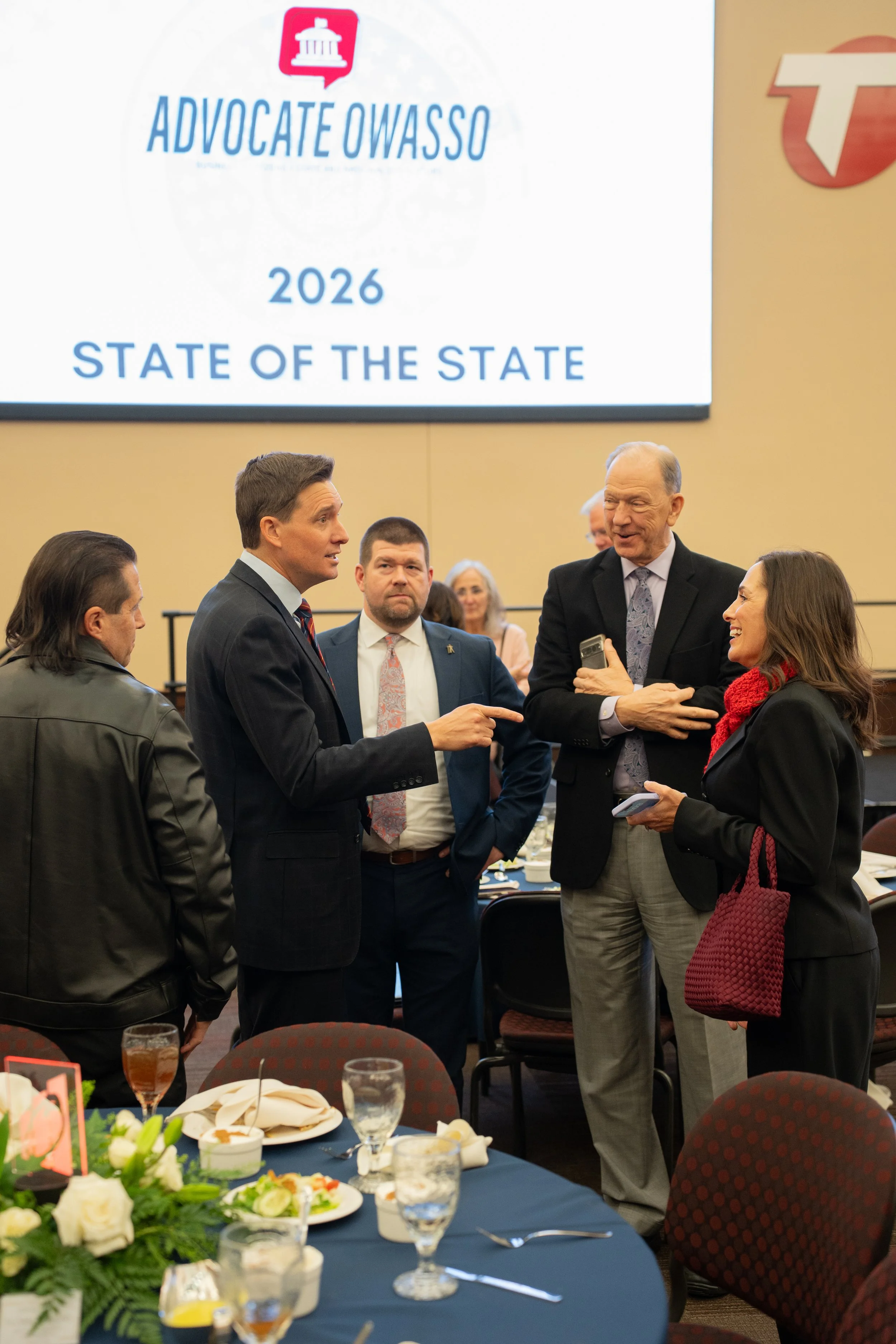 Group of five people engaged in conversation at a formal event, with a screen in the background displaying "Advocate Owasso 2026 State of the State." Tables set with plates, glasses, and a floral centerpiece are visible in the foreground.