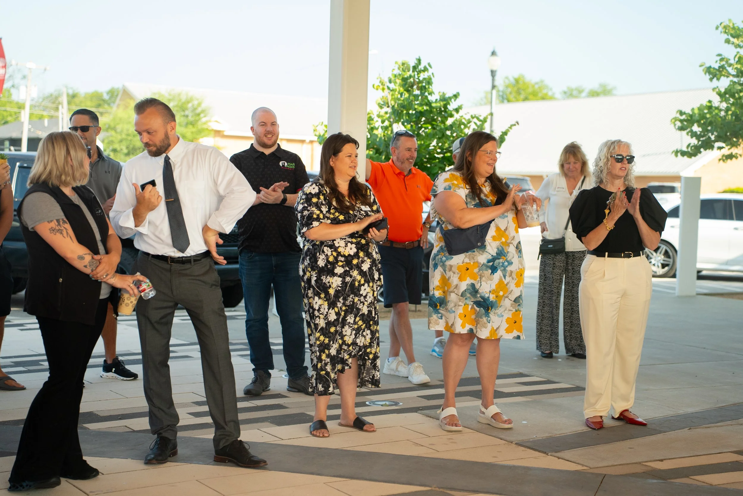 Group of casually dressed people standing outdoors, some clapping, watching an event or person off-camera.