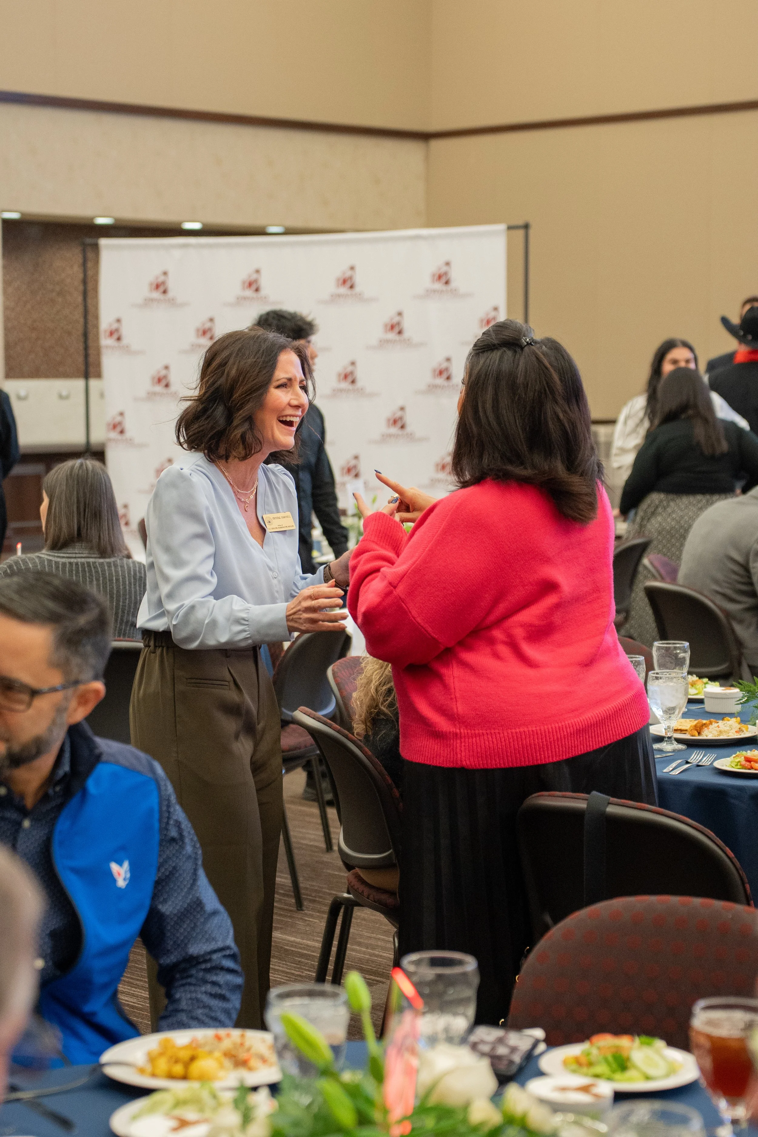 Two women engaged in conversation at a formal event, with the woman on the left wearing a light blue blouse and the woman on the right wearing a red sweater. The event appears to be a gathering or conference, with several attendees seated at round tables with plates of food and glasses of water.