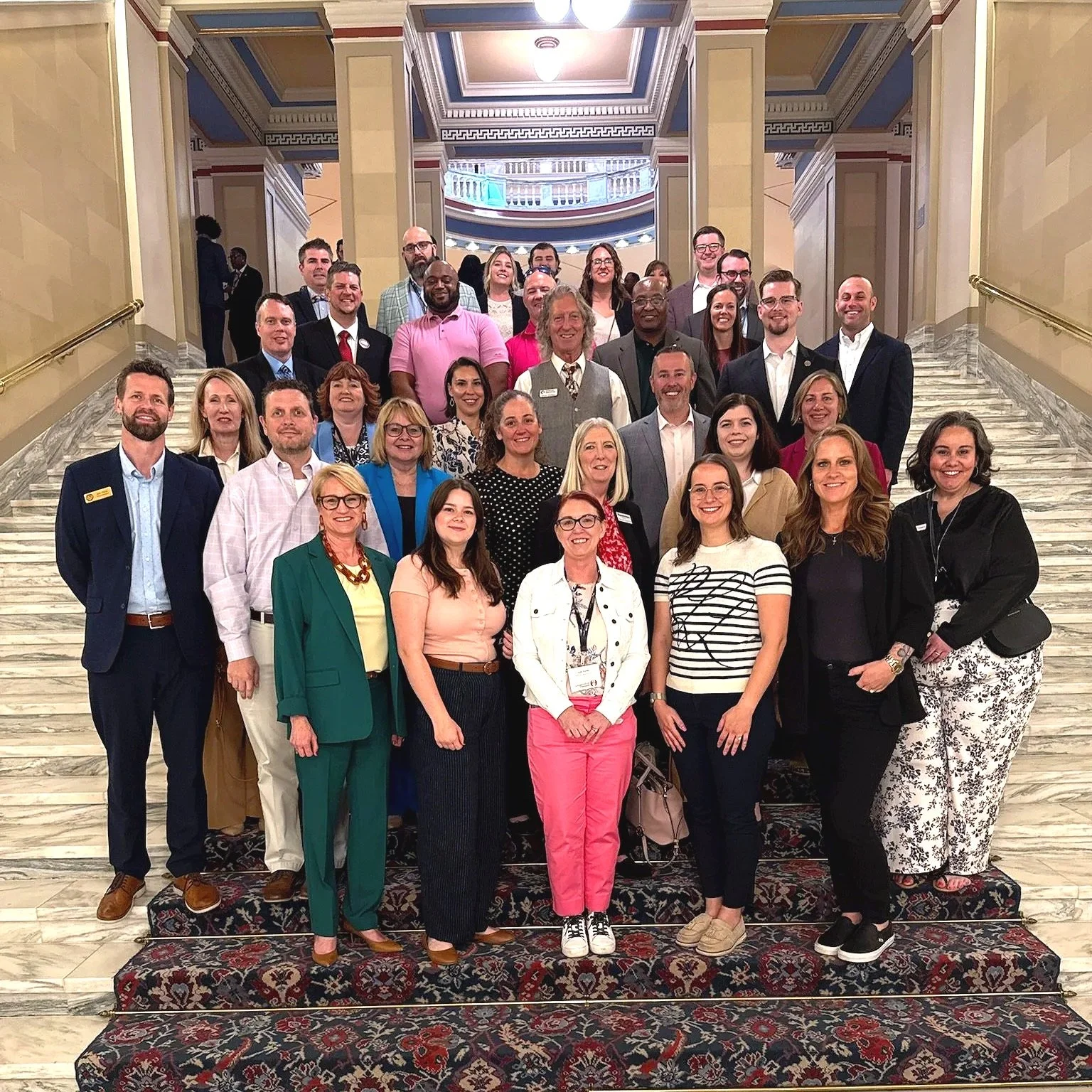 Group of diverse professionally dressed people standing on marble stairs in a grand hallway with ornate details and a balcony in the background.