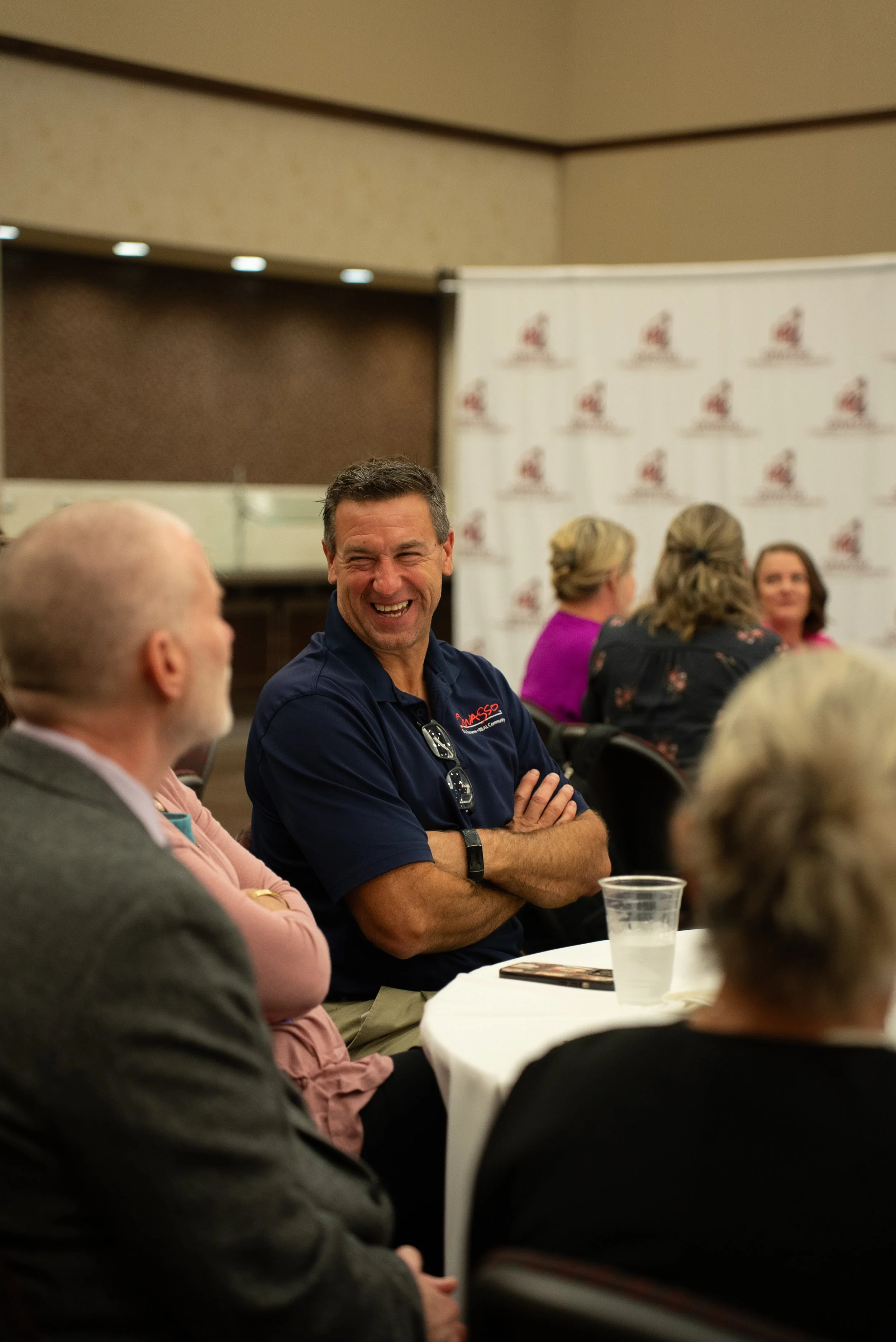 Group of people sitting at round tables in a meeting room, with a man in a navy polo shirt laughing in the center.