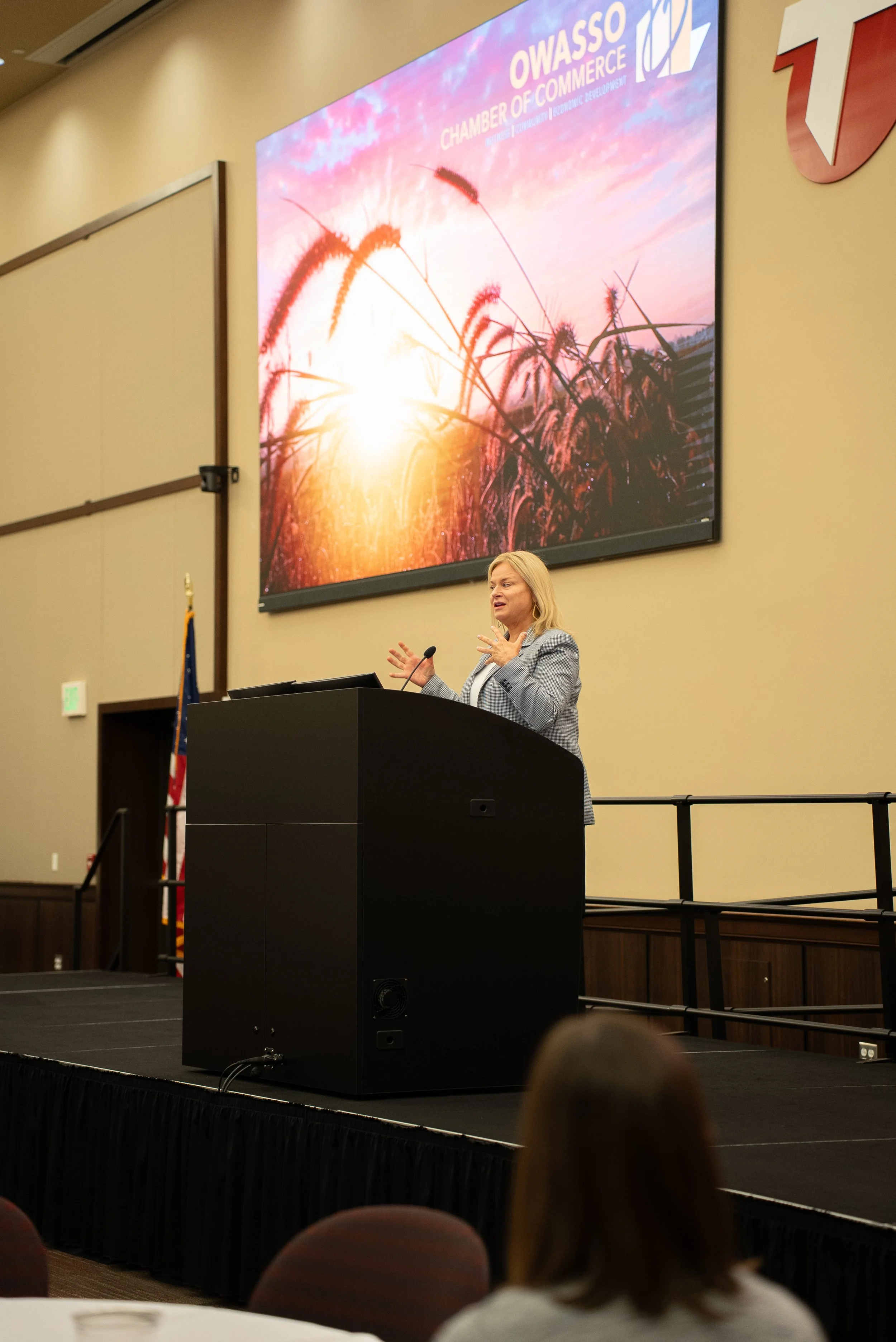 A woman giving a presentation at a conference with a large screen behind her displaying a sunset and wheat field image with the words 'OWASSO CHAMBER OF COMMERCE' at the top.