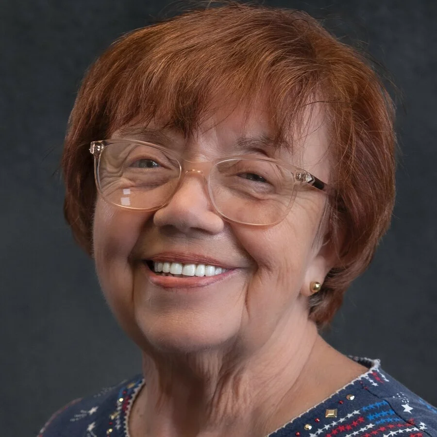 A smiling elderly woman with short red hair, wearing glasses, earrings, and a colorful top, against a dark background.