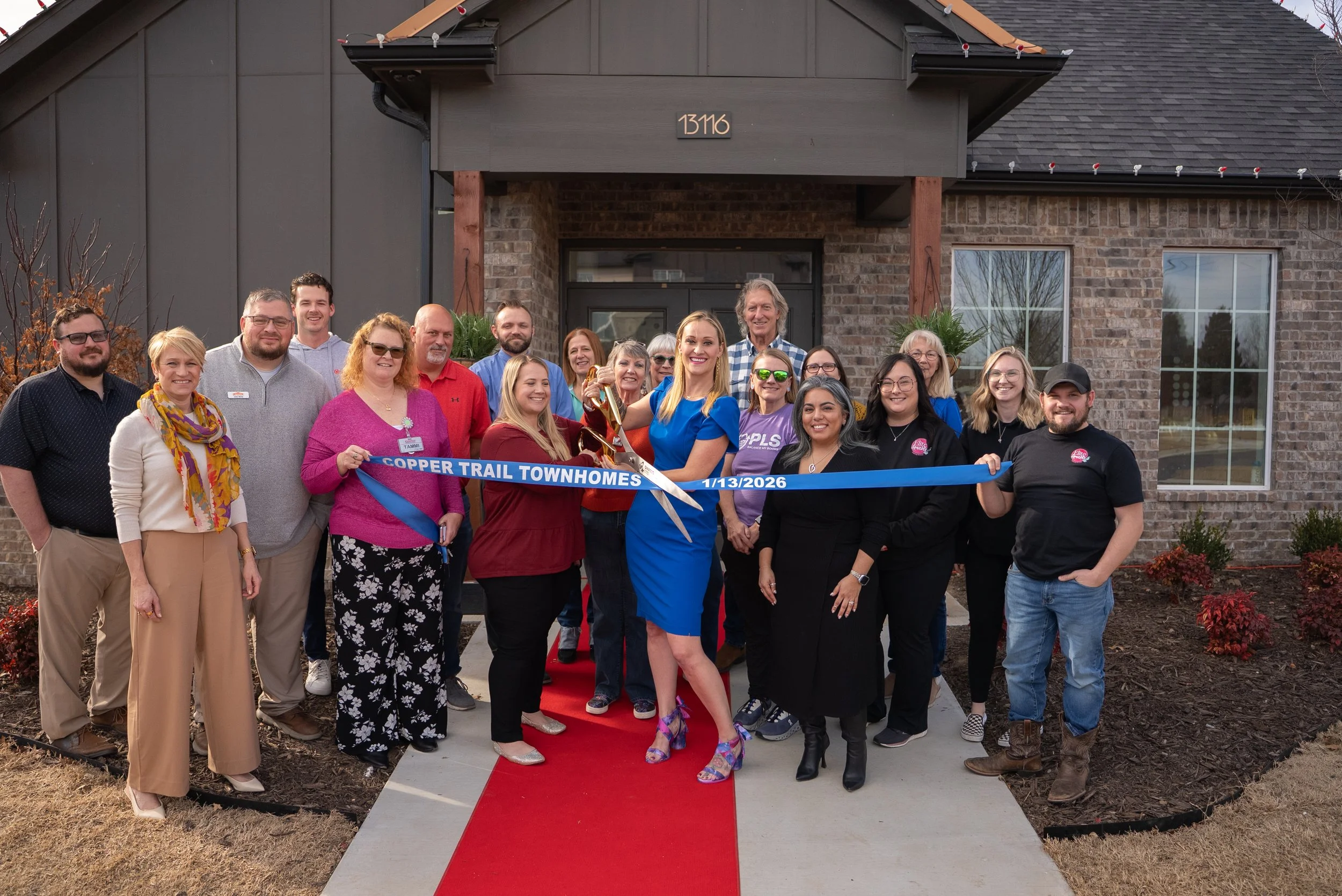 Group of people celebrating the grand opening of Copper Trail Townhomes, holding a ribbon in front of a newly constructed building with a red carpet on the sidewalk, date 1/13/2026.