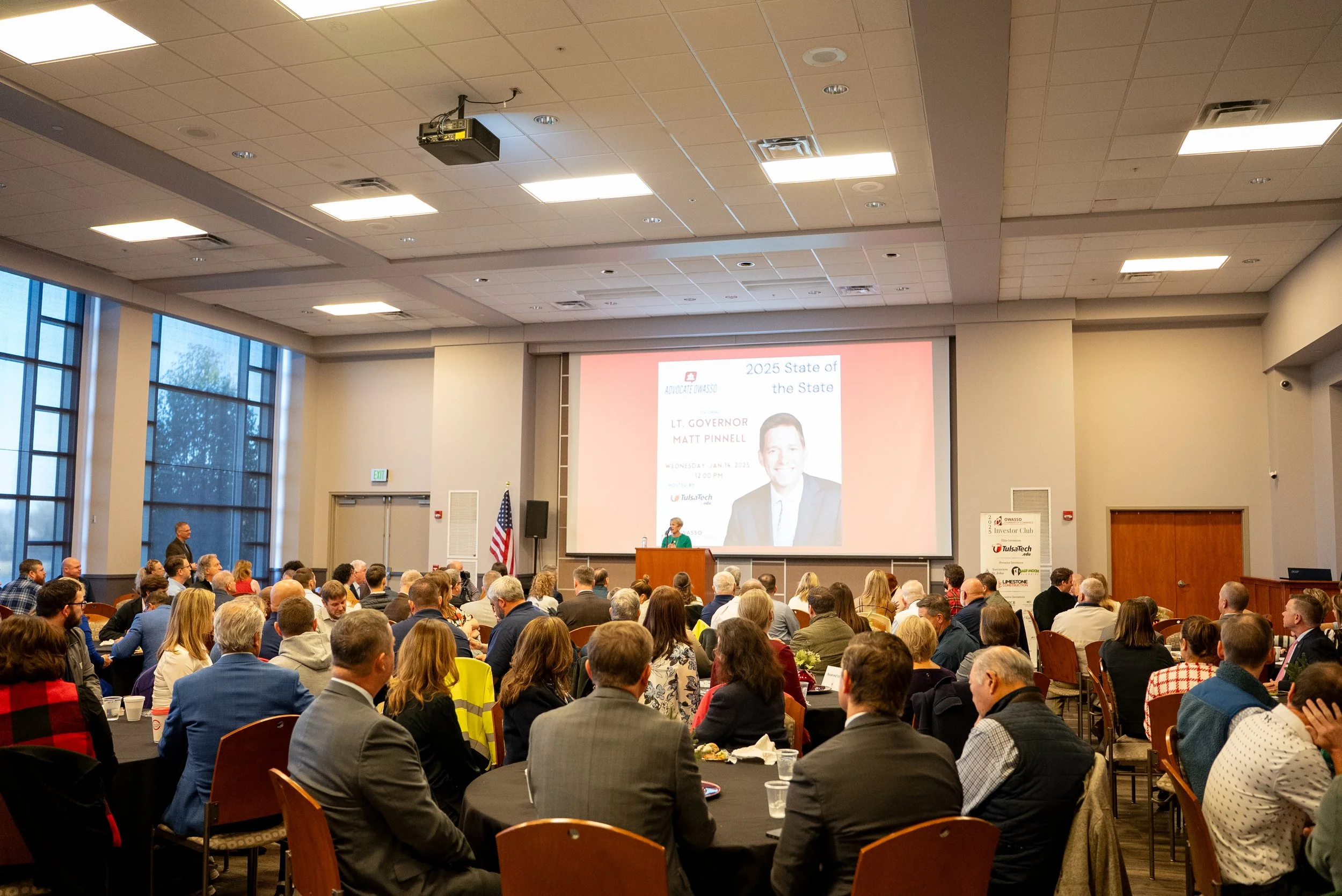 A large conference room filled with people attending a presentation or speech. A speaker is standing at a podium on a stage in front of a large screen displaying a slide with a photo of a man and text about a 2025 State of the State event, Lt. Governor Matt Pinnell, on January 14, 2023. The room has large windows on the left, and American flags, with people seated at round tables.