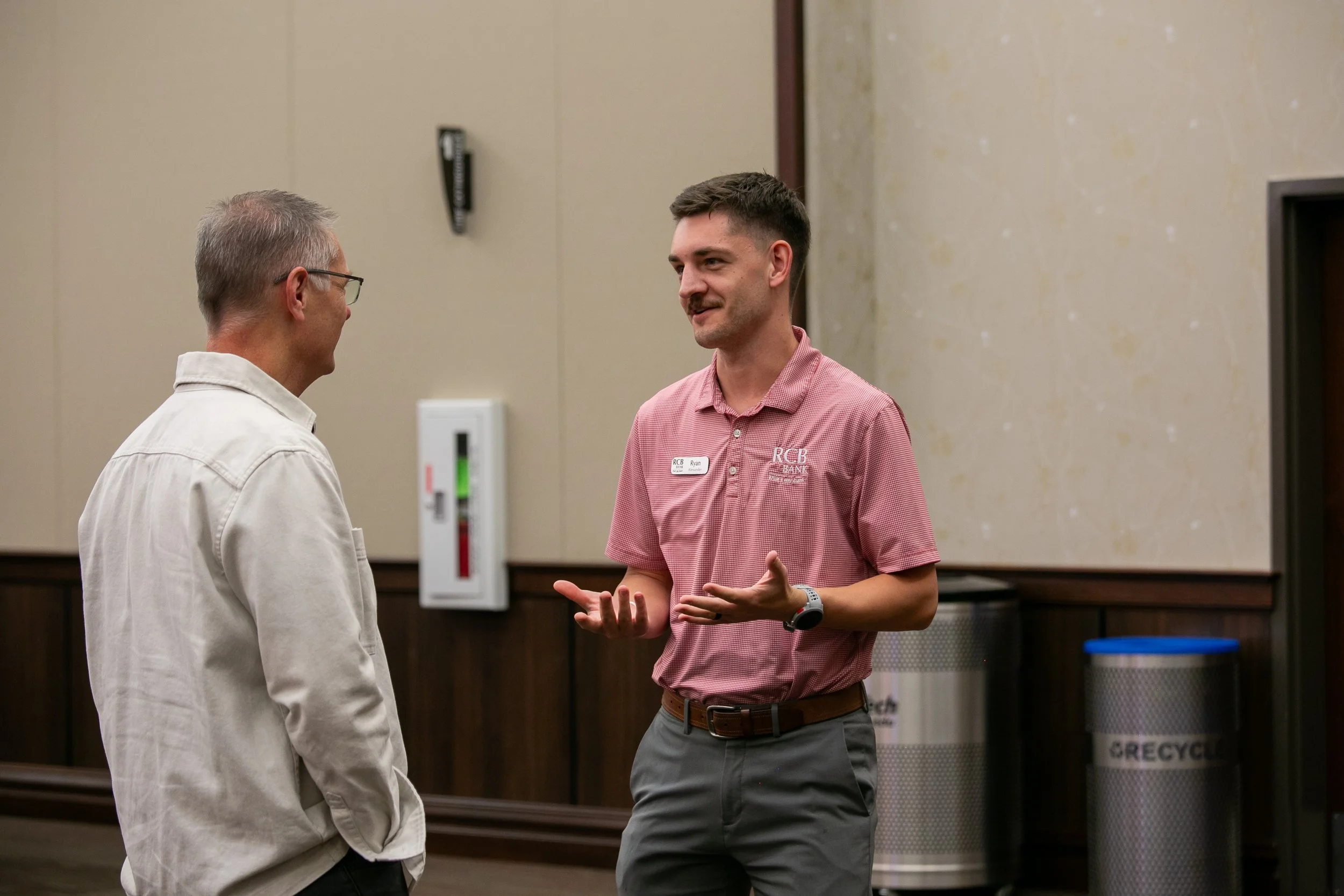 Two men are talking in a room, one wearing a beige jacket and glasses, the other in a red short-sleeve shirt with a name tag, gestures with his hands while speaking.
