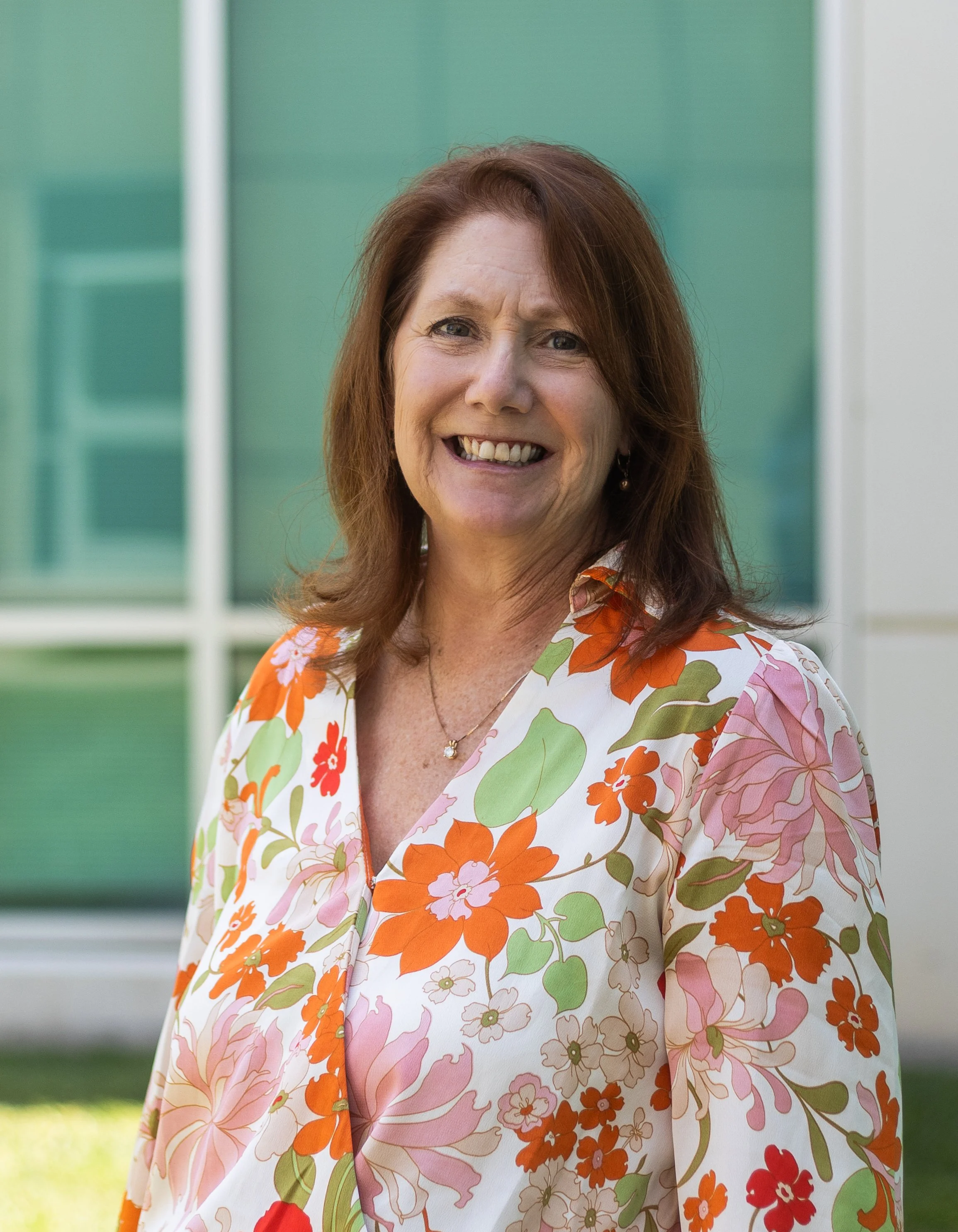 A middle-aged woman with shoulder-length brown hair, smiling outdoors in front of a building with green and white windows, wearing a floral patterned blouse with orange, pink, and green flowers.