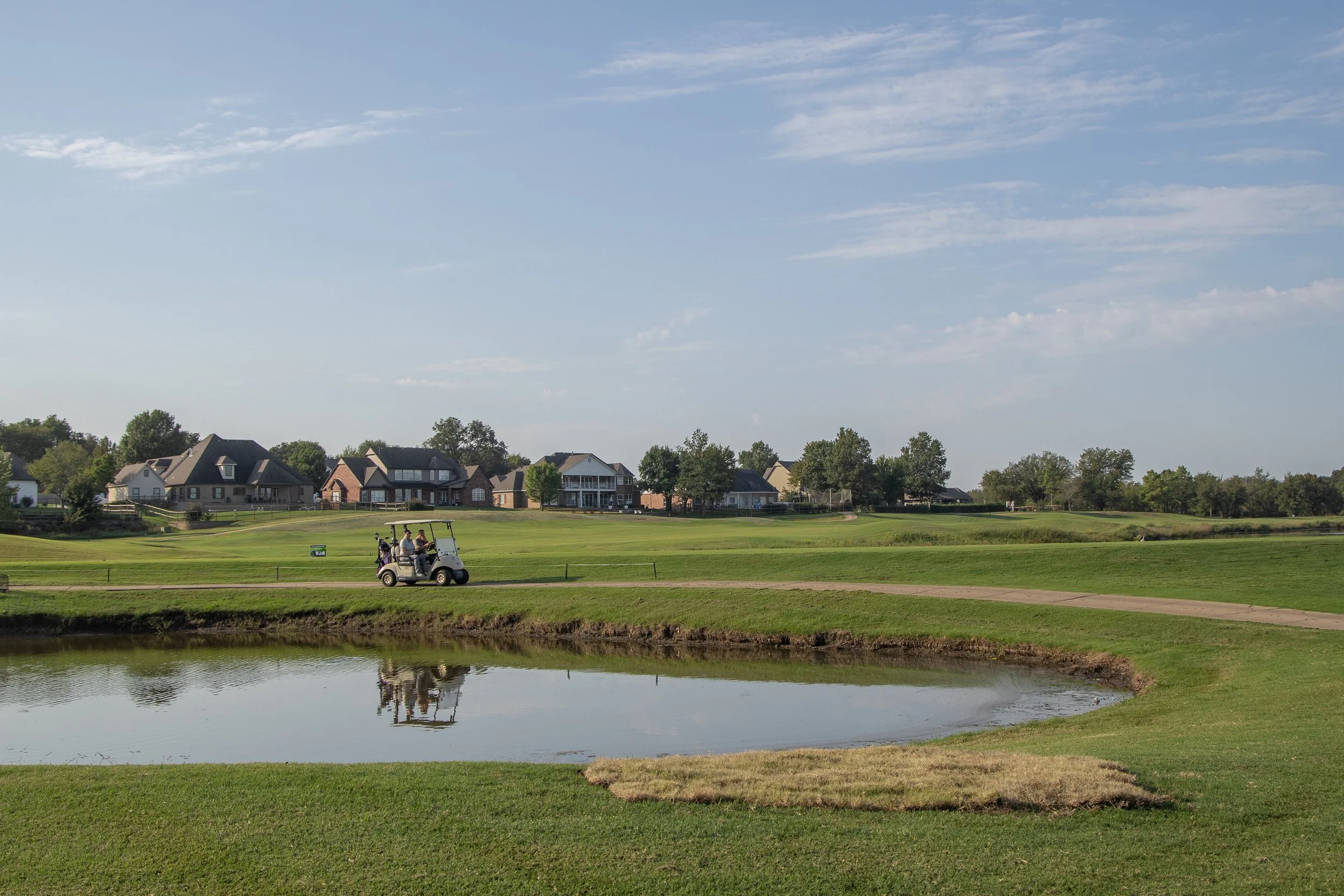 A golf course with a small pond, a golf cart with players, and residential houses in the background under a partly cloudy sky.