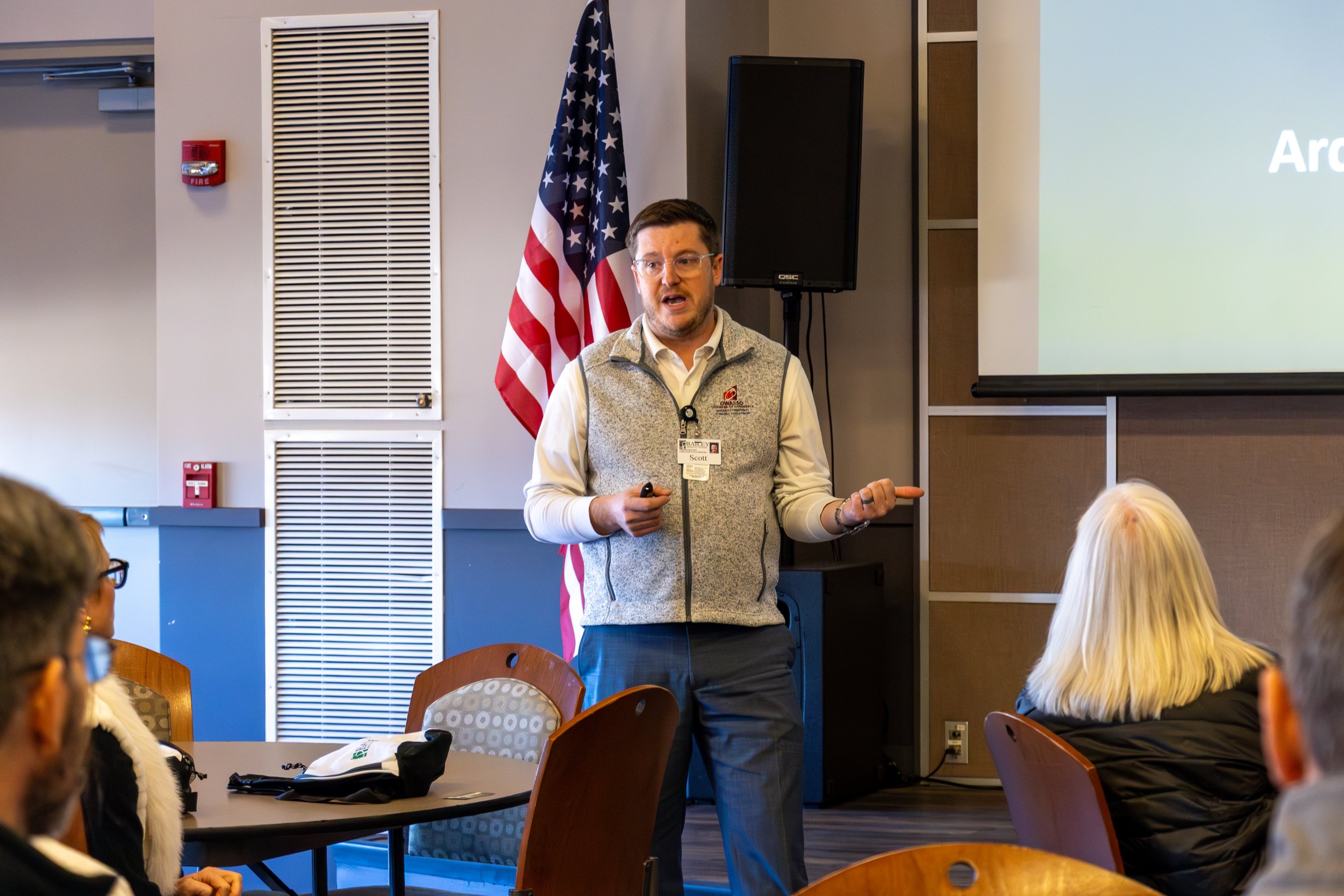 A man with glasses and a beard giving a presentation in a conference room with American flag in the background. The man is wearing a gray vest over a white shirt and is holding a remote in his right hand. There are people seated at tables facing him, including a woman with blonde hair and another person with glasses. A presentation slide is projected on the wall to the right.