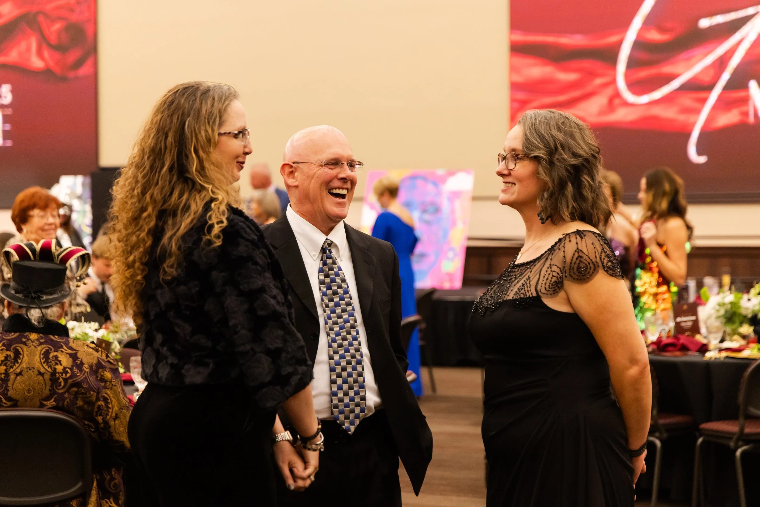 Three people are smiling and talking at a celebratory event. Two women and a man, with the man wearing a suit and tie, and the women wearing black dresses, are standing and facing each other. There are tables with decorations and other people in the background.