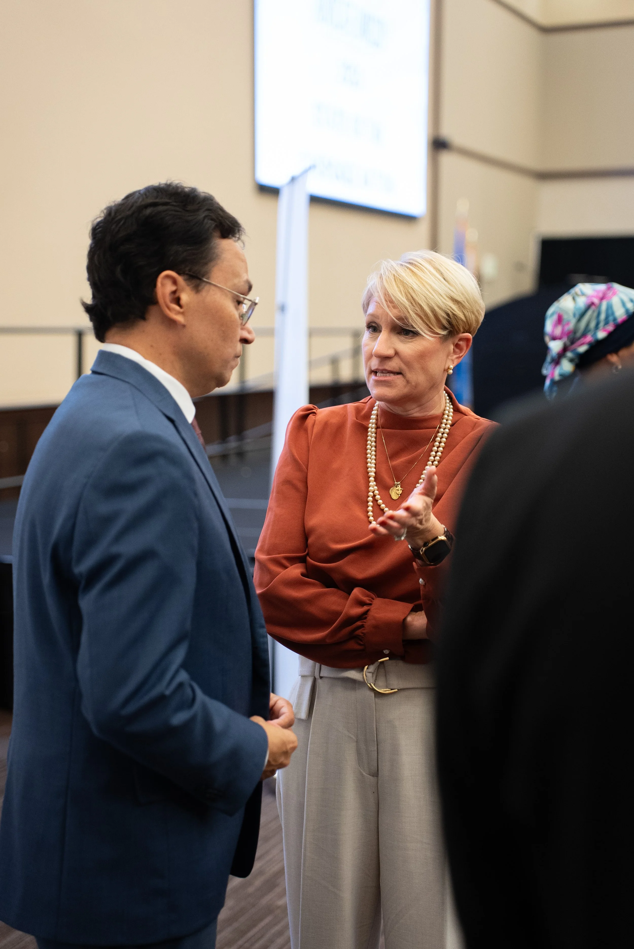 A woman with short blonde hair, wearing a rust-colored blouse, cream-colored pants, and pearl jewelry, is speaking and gesturing towards a man with dark hair, glasses, and a blue suit, in a professional setting.