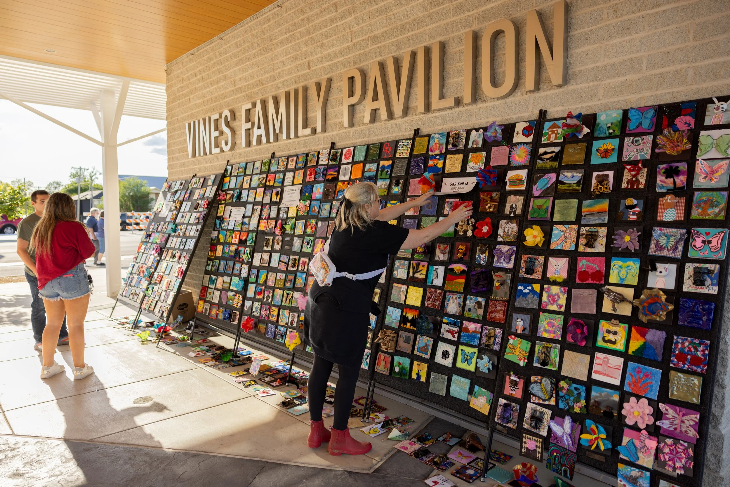 People browsing and selling artwork at an outdoor art display outside a building labeled "Vines Family Pavilion" with a large collage of small colorful art pieces on display.
