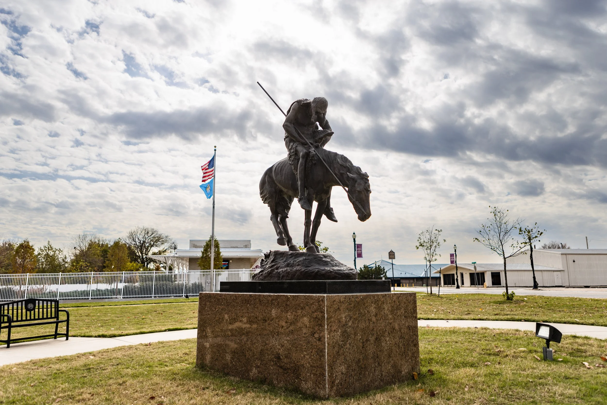 Bronze statue of a Native American man on horseback, holding a spear, in a park with flagpoles, trees, and buildings in the background under a cloudy sky.